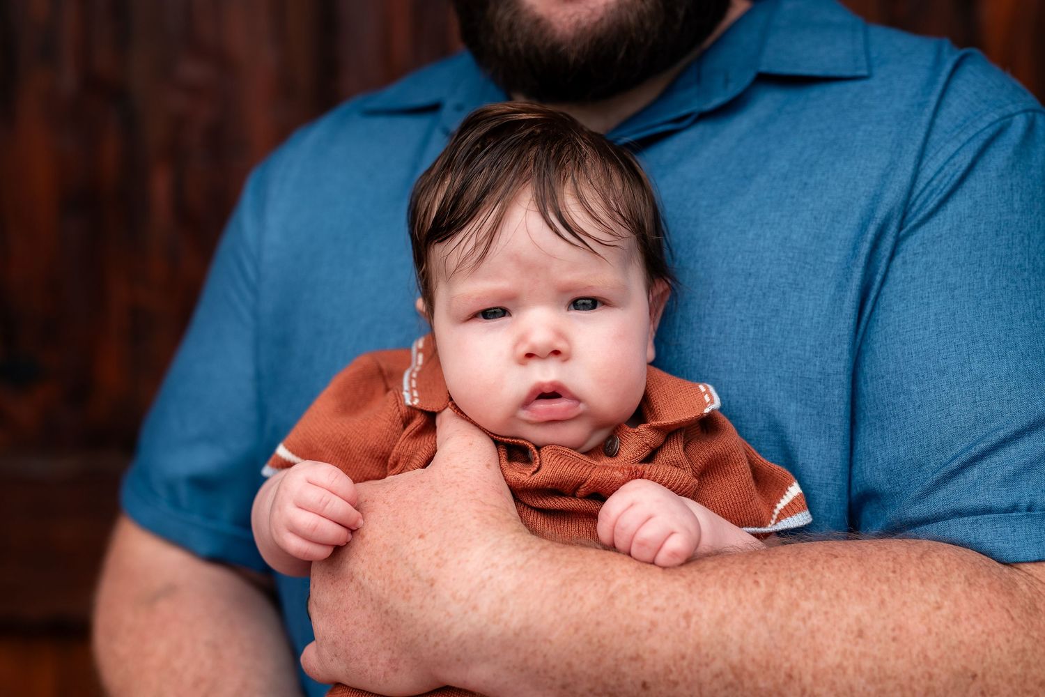 A bearded father in a blue polo shirt gently holds his newborn baby dressed in a rust-colored onesie.