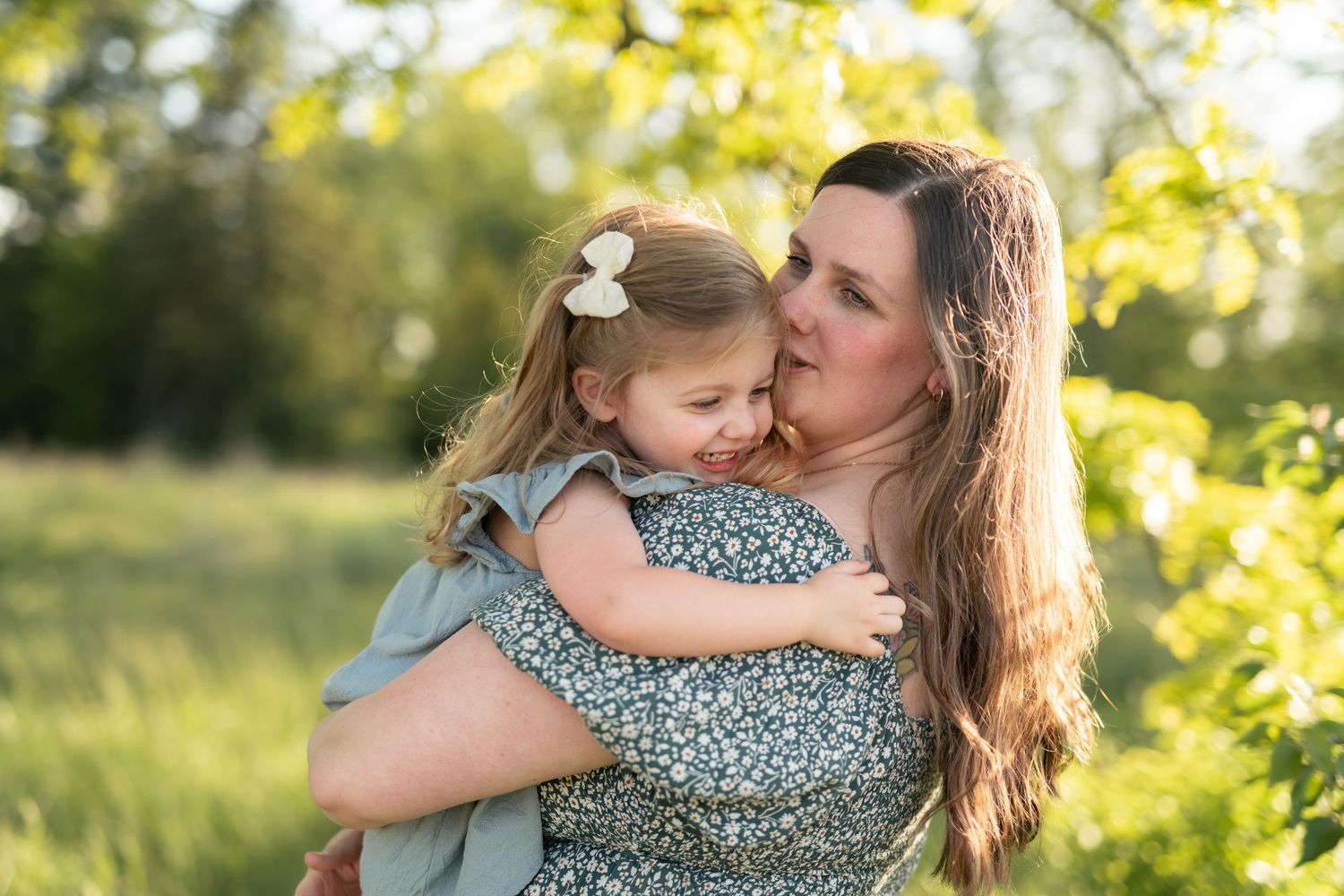 A mother tenderly kisses her young daughter outdoors in a sunlit green park setting.