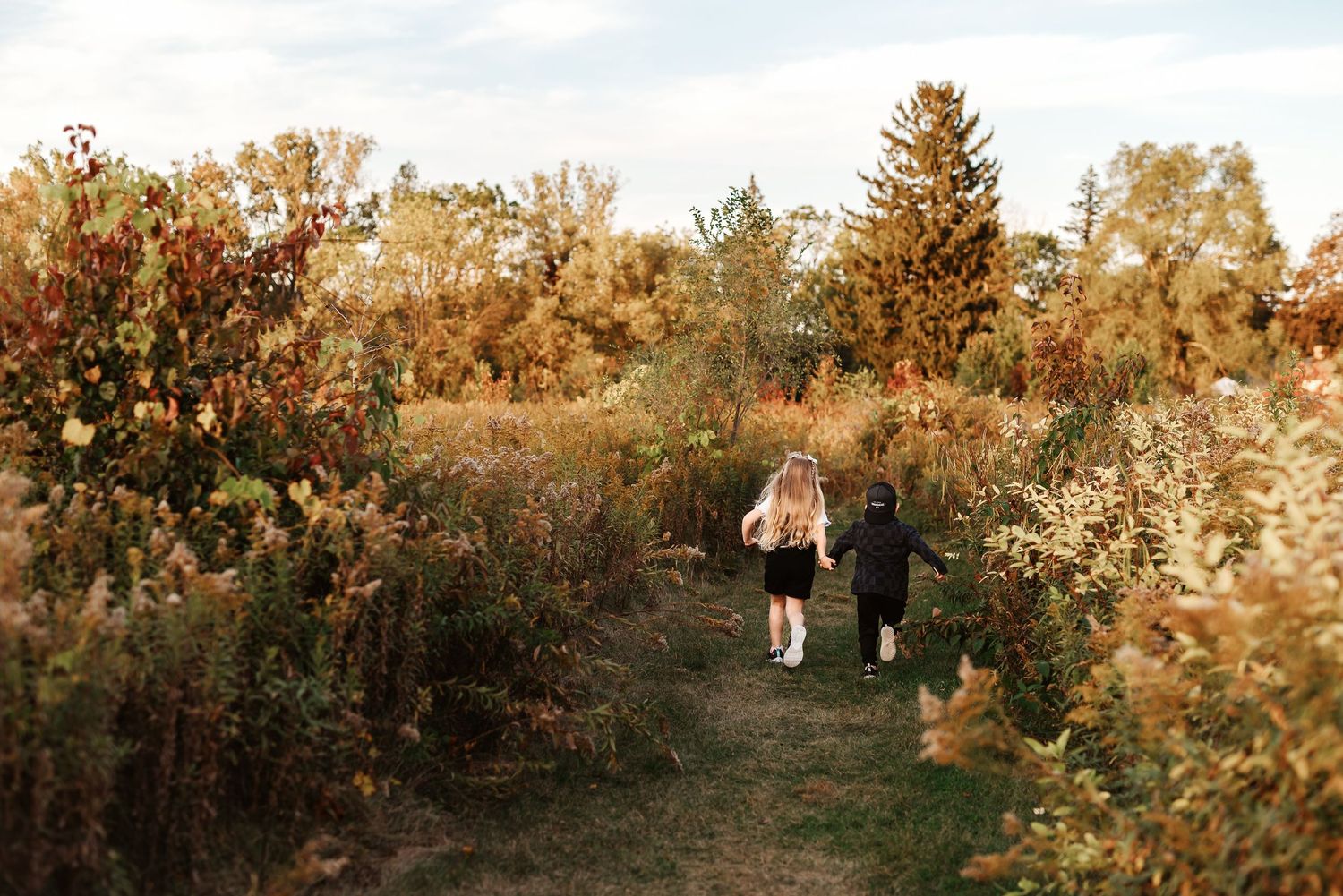 Two children walk hand-in-hand through a scenic autumn trail surrounded by colorful fall foliage.