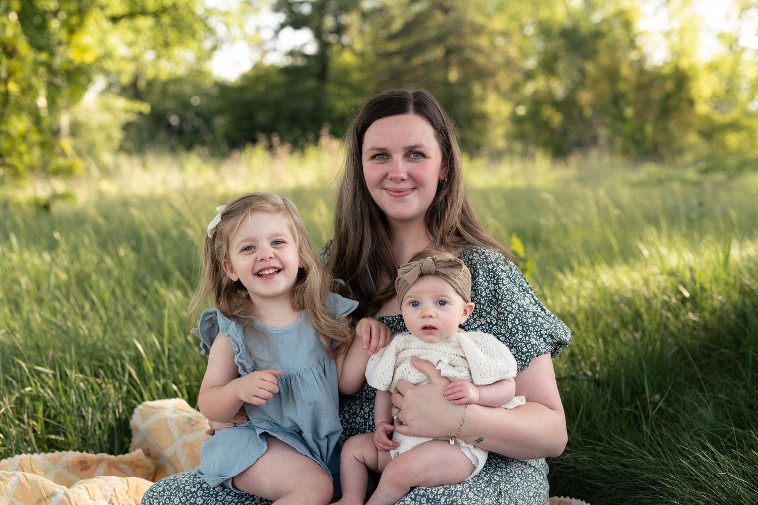 A mother sits outdoors in a green field with her two young daughters, smiling warmly during a family portrait session.