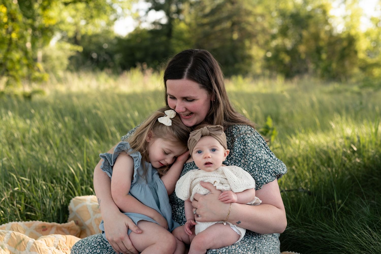 A mother sits in a green field holding two young children, smiling warmly in soft golden sunlight.