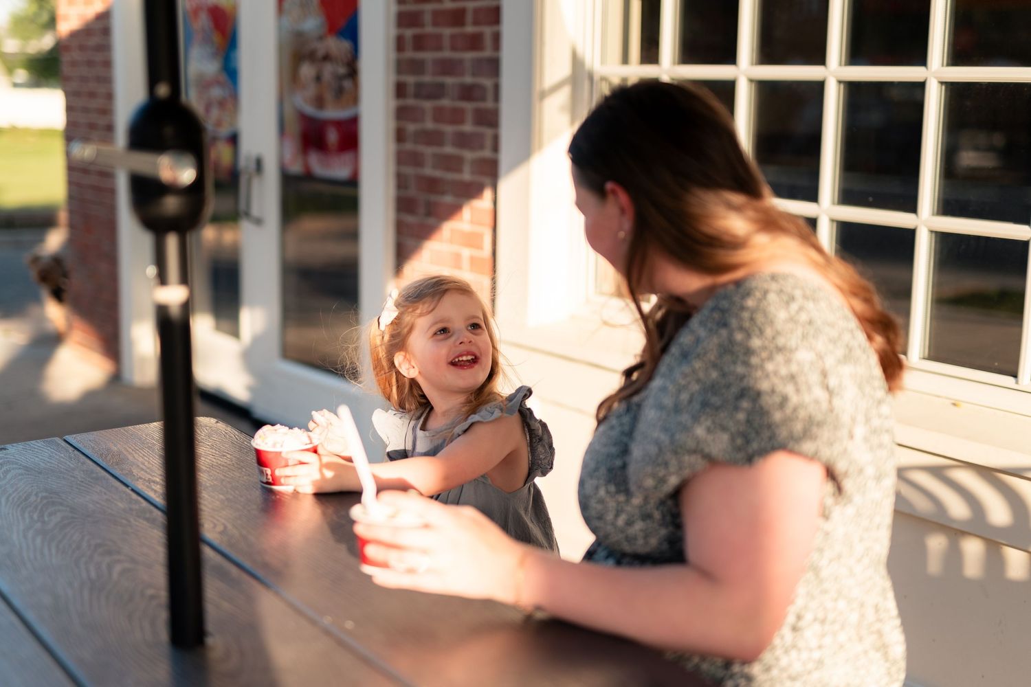 A mother and young daughter share a joyful moment outdoors at a table with red cups in warm sunlight.