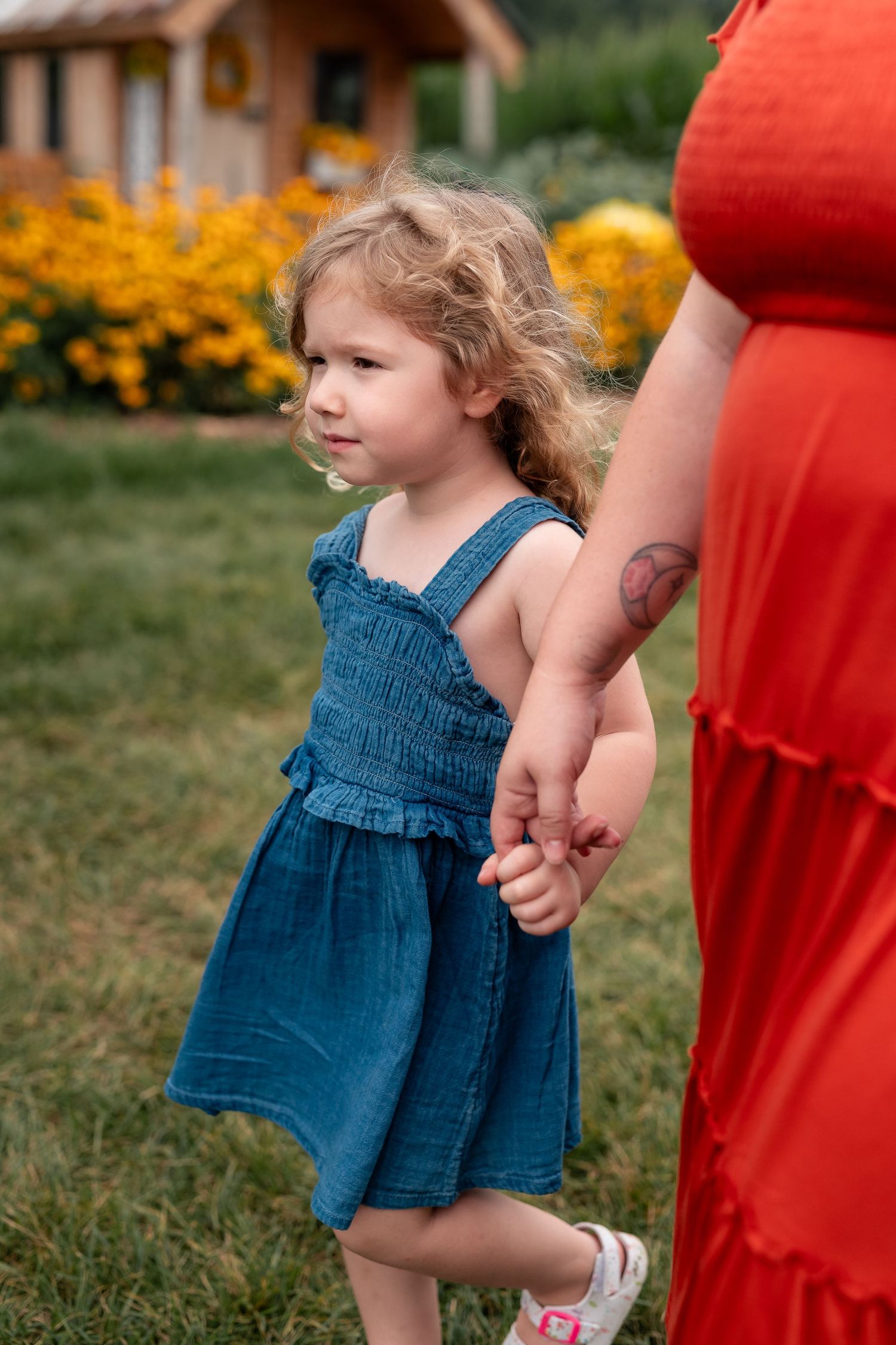 A young girl in a blue denim dress holds her mother's hand in a garden with yellow flowers.