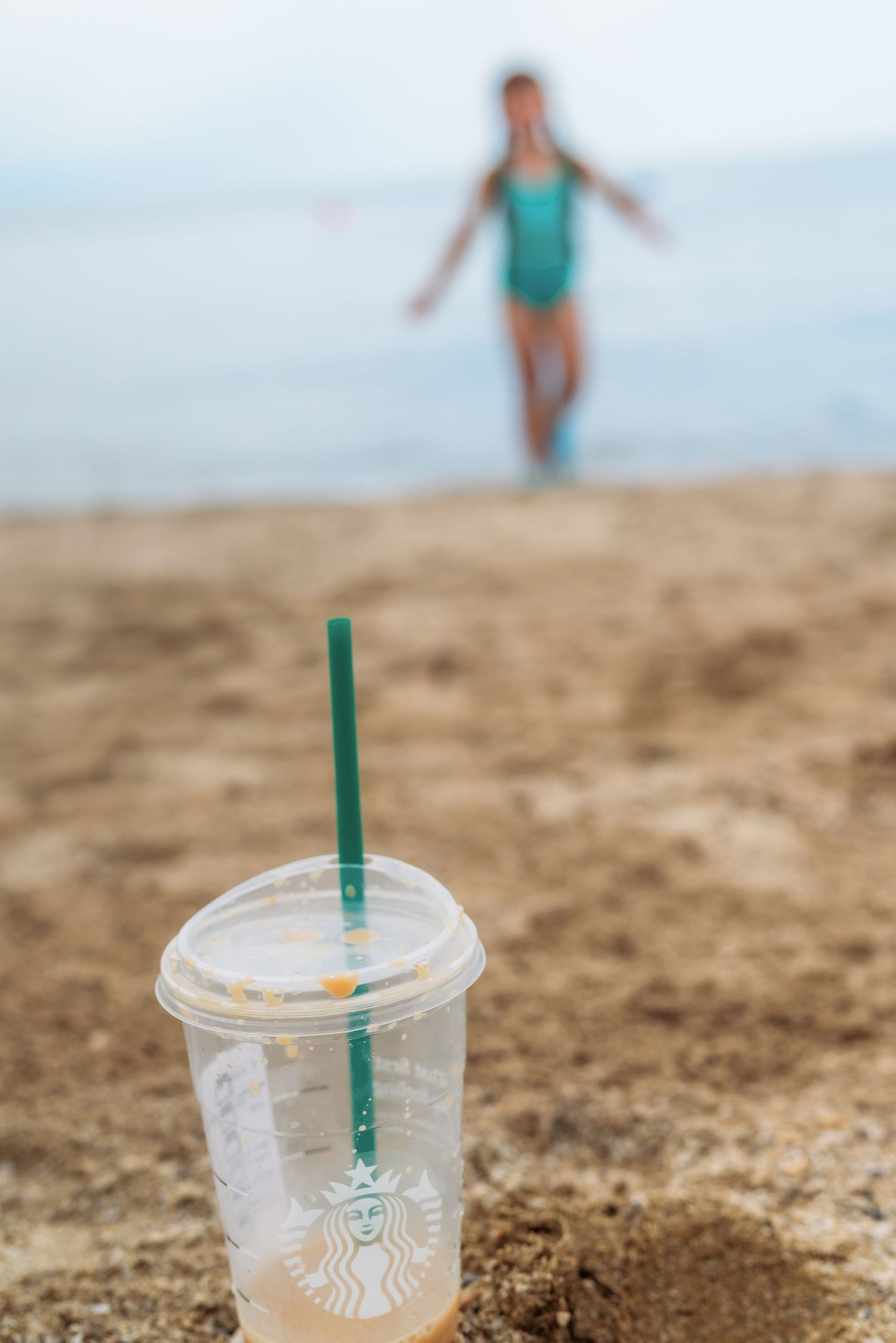 Empty Starbucks plastic cups with green straws in the sand with blurred people playing in ocean waves in background.