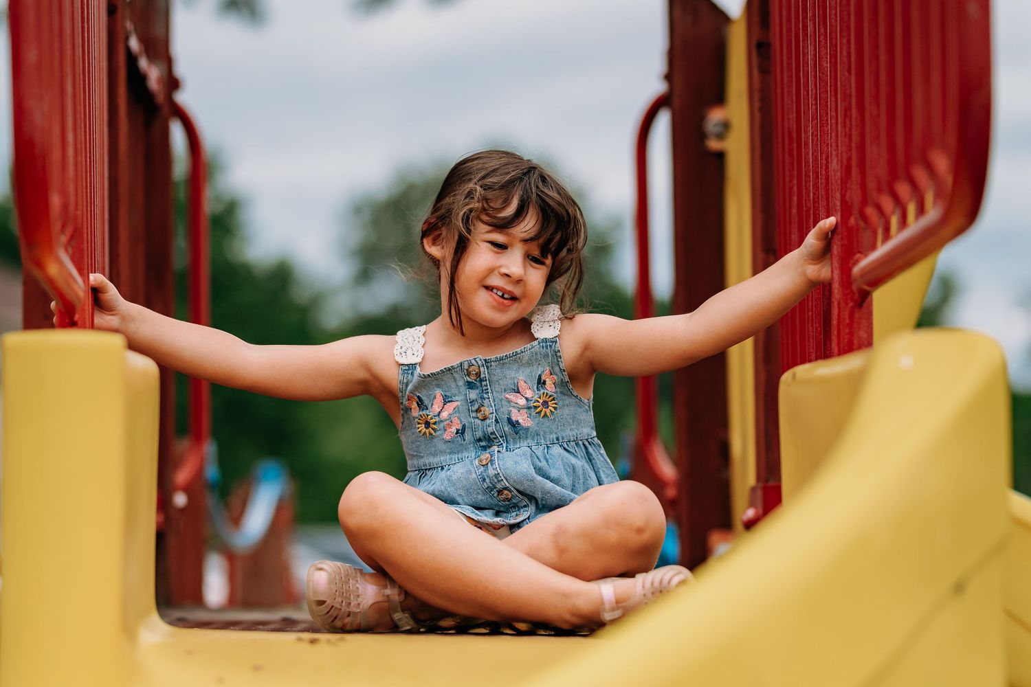 Child playing on a yellow playground slide with arms outstretched, wearing a floral patterned outfit.