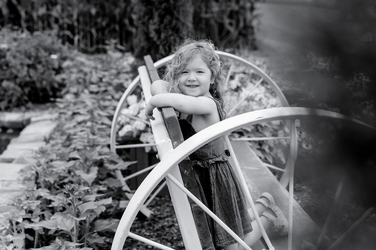 Happy young girl smiling while leaning on decorative white wheel arch in lush garden.