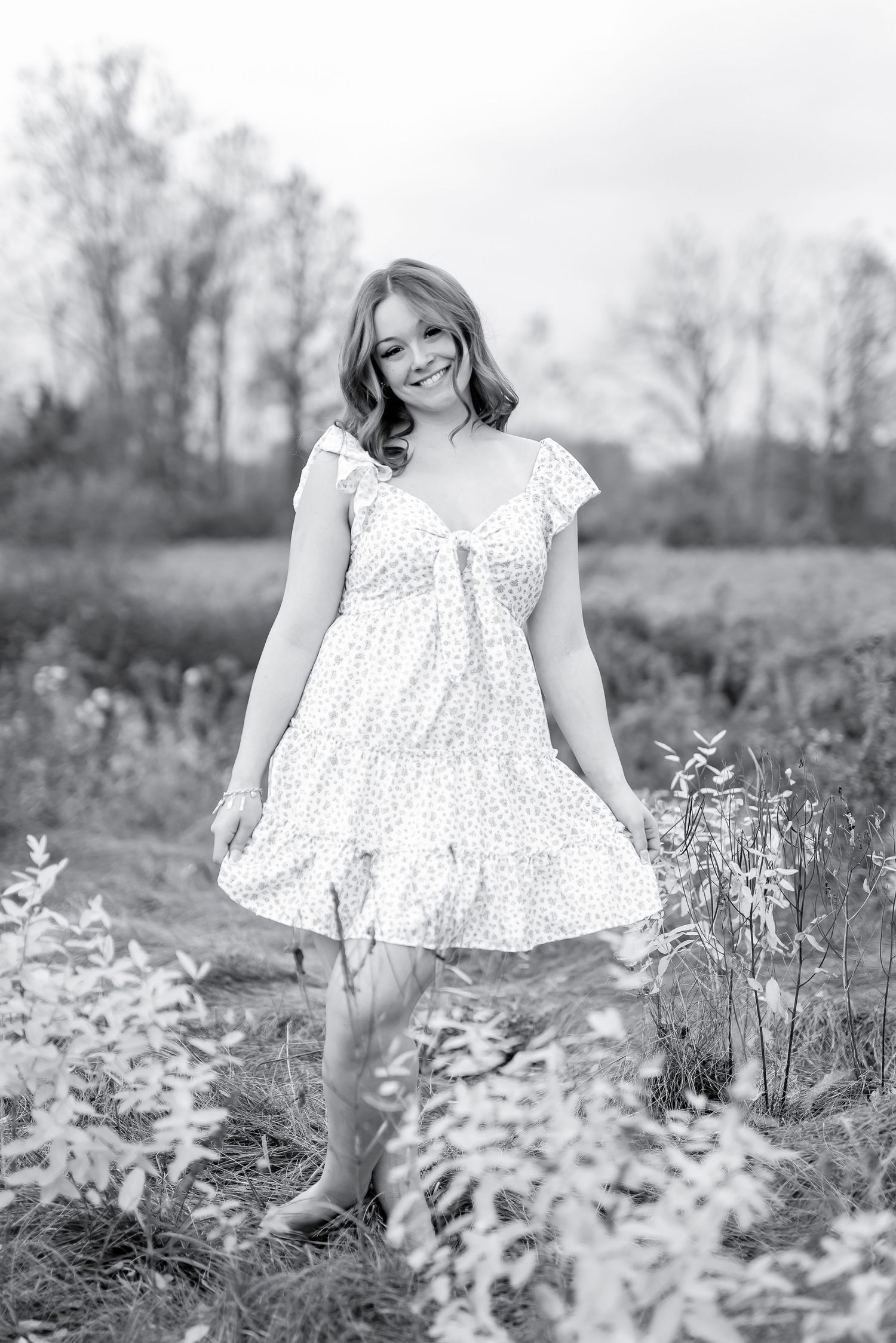Black and white photo of smiling woman in floral dress posing in sunny meadow.