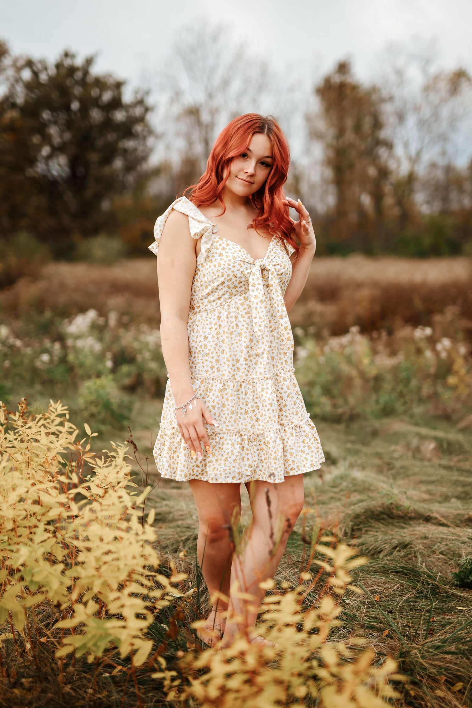 Red-haired woman in white floral sundress standing among golden autumn plants in a field.