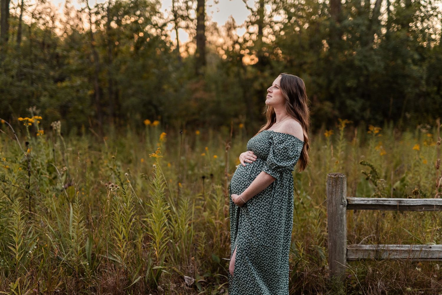 Pregnant woman in green floral off-shoulder dress standing in golden hour field near wooden fence.
