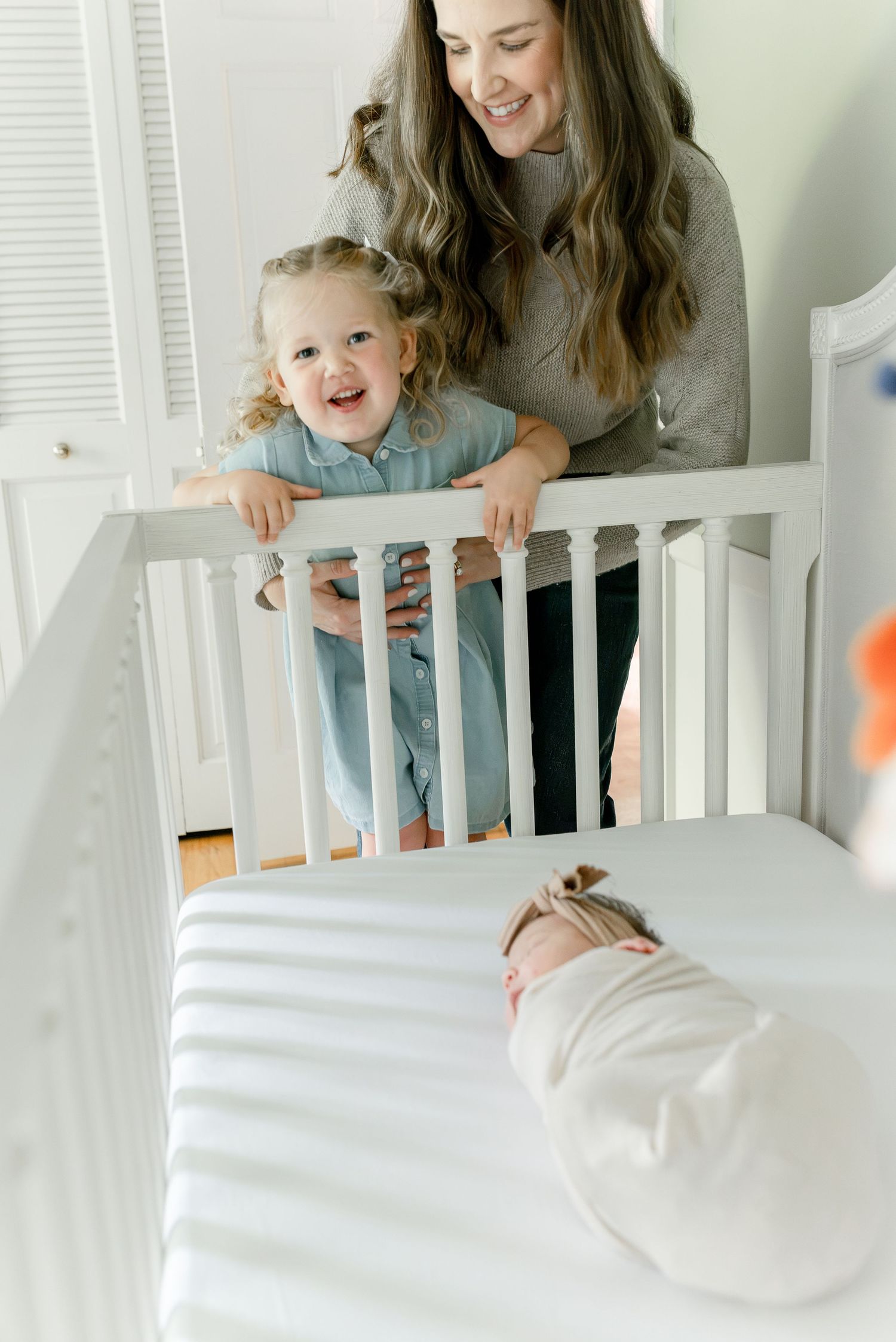 Smiling mother and excited toddler girl looking at newborn baby lying in white crib.