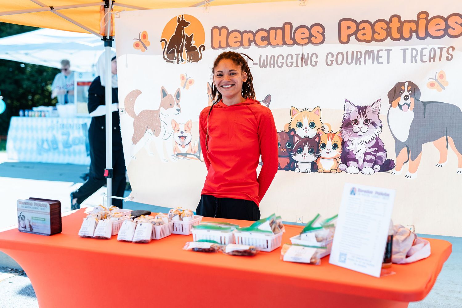 Young girl smiling at Hercules Pastries gourmet pet treats vendor booth at an outdoor market event.