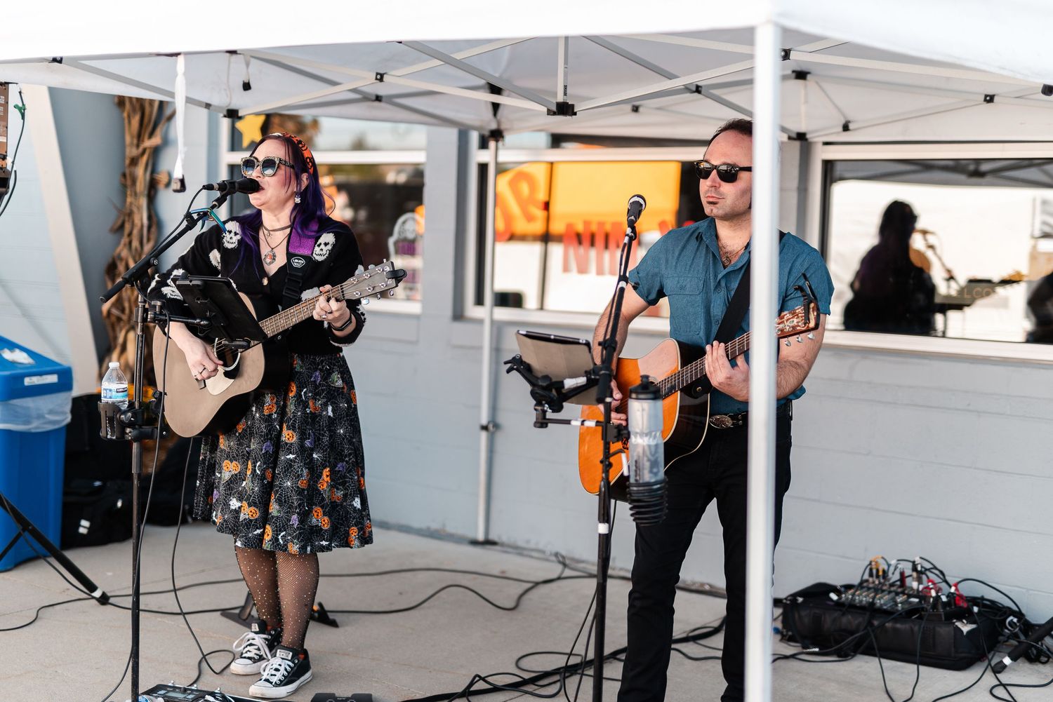 Two musicians performing live under a white canopy tent, woman playing guitar in floral dress and man with violin.