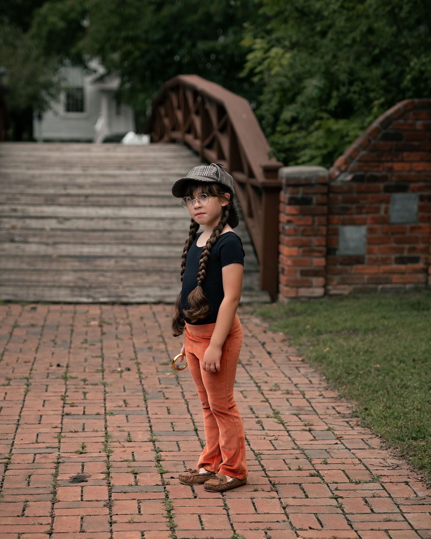 Young girl in orange pants and newsboy cap stands on brick path near ornate metal bridge.