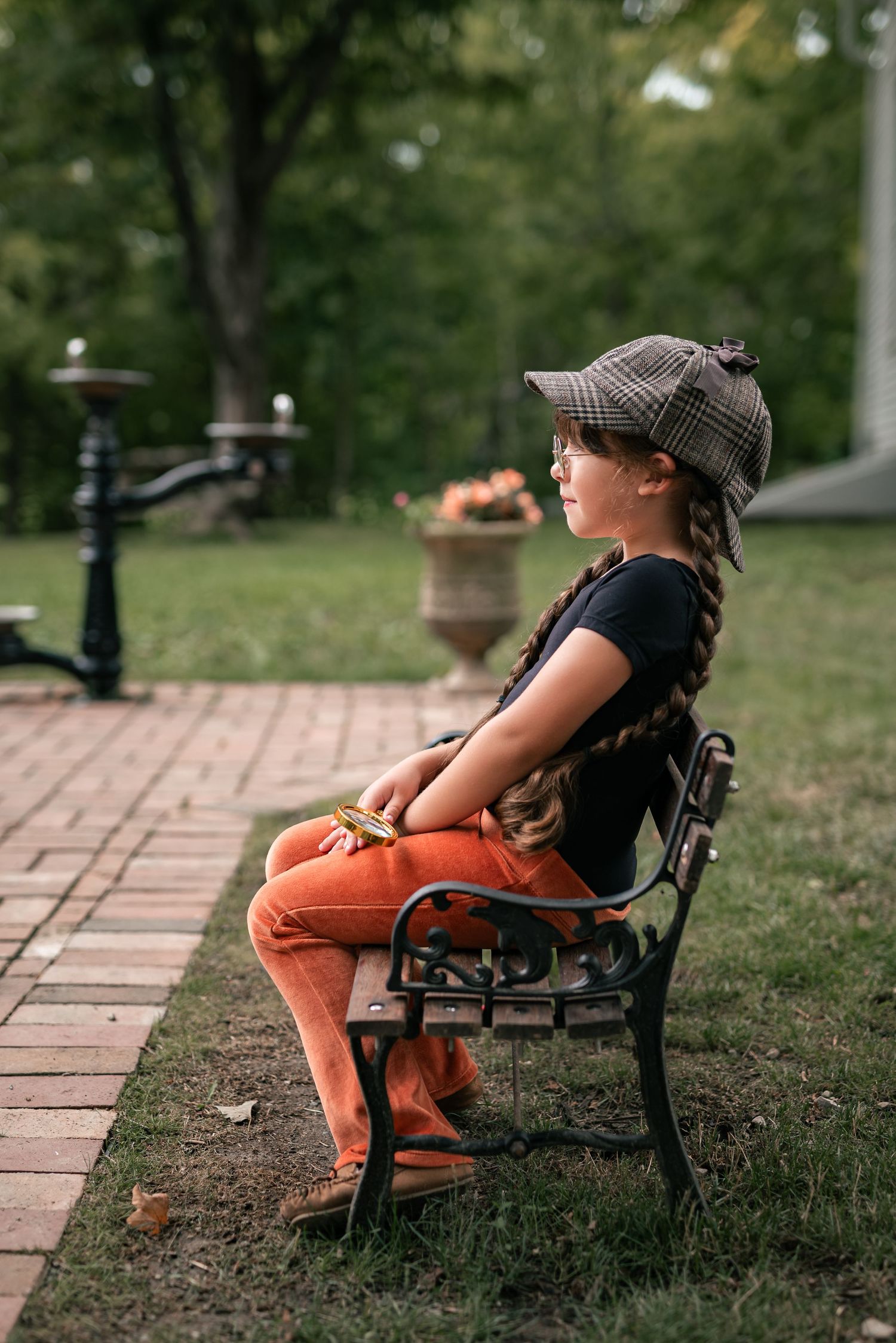 Young girl in plaid cap and orange pants sitting on ornate park bench, gazing thoughtfully at lush green garden.