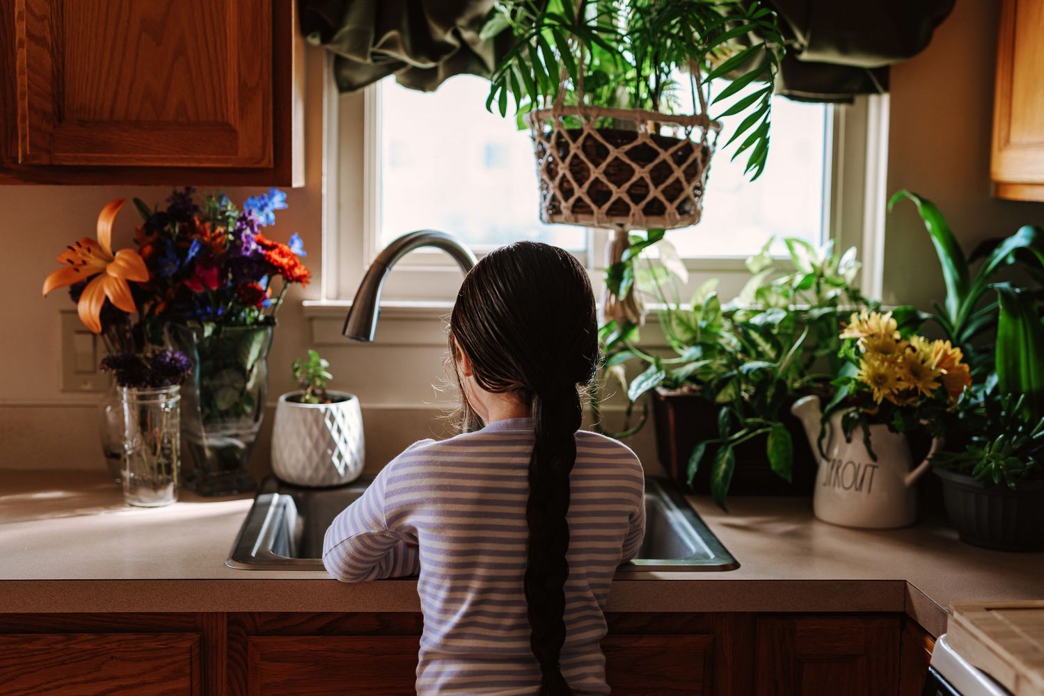 Someone in a striped shirt washes dishes at a kitchen sink surrounded by houseplants and floral arrangements.