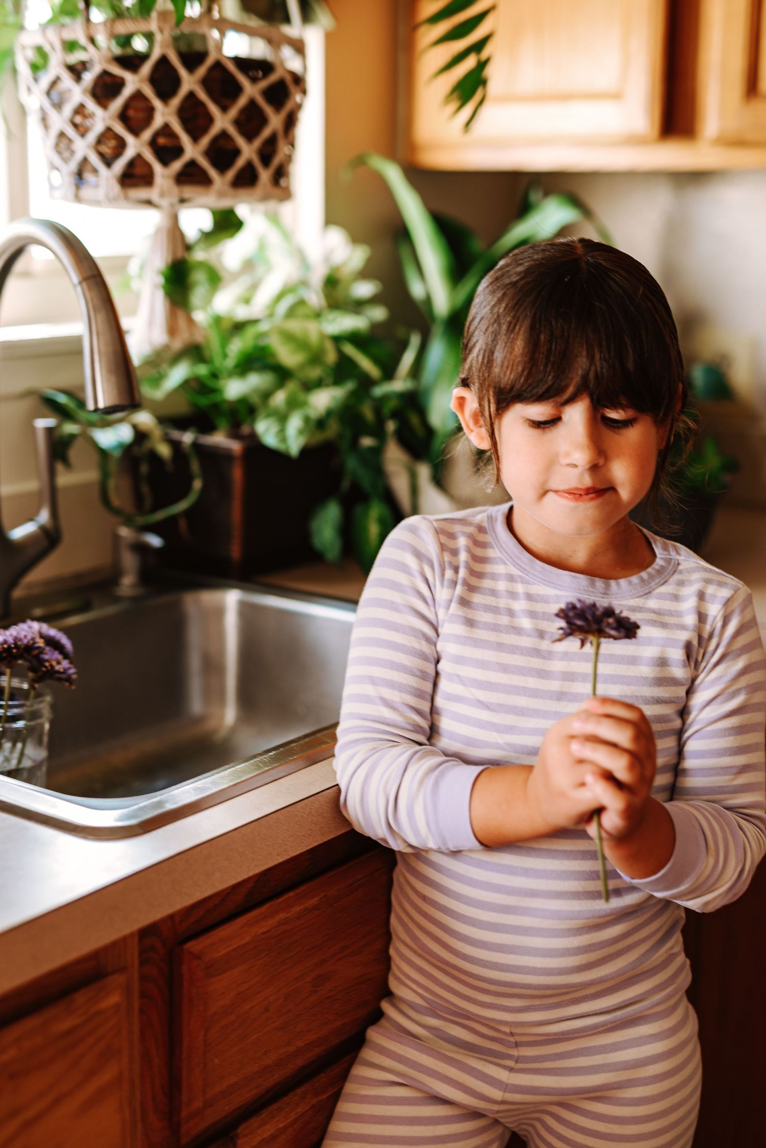 A child in a striped shirt stands at a kitchen counter holding and examining a small purple flower.