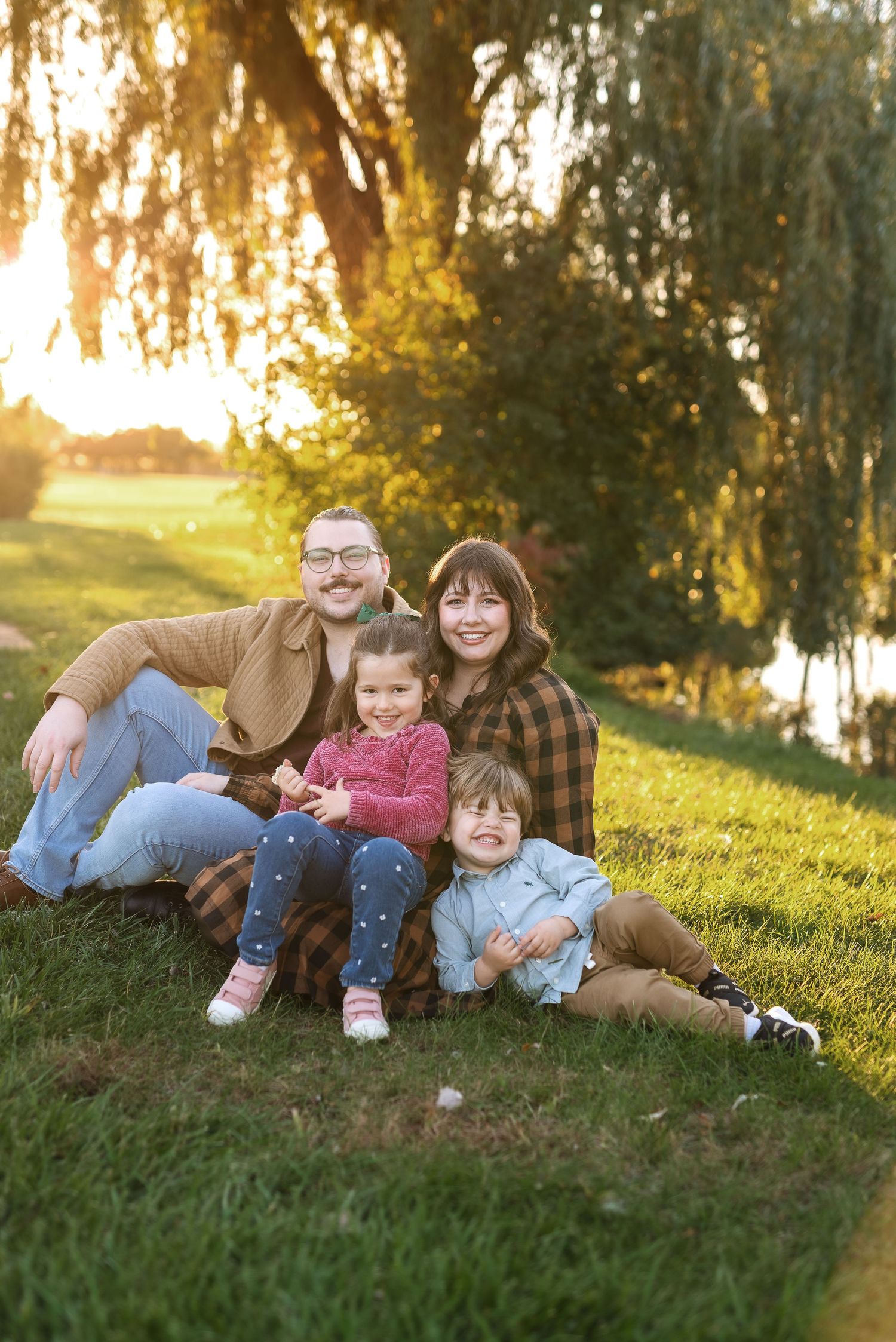 Family lounges together on grassy hillside during golden hour sunset in casual outdoor portrait session.
