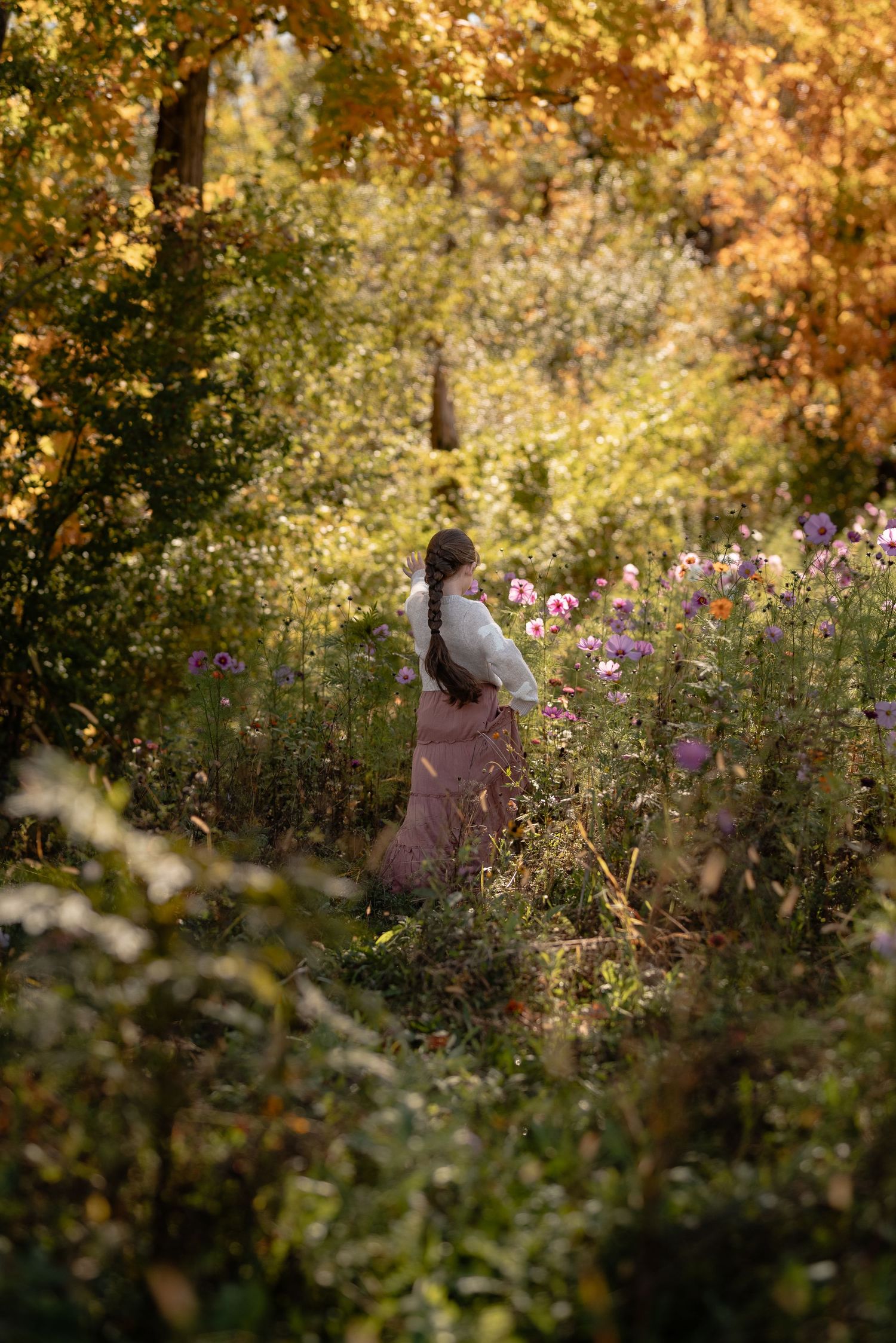 Woman with a long braid in a white blouse and pink skirt stands among purple wildflowers in a golden autumn forest.