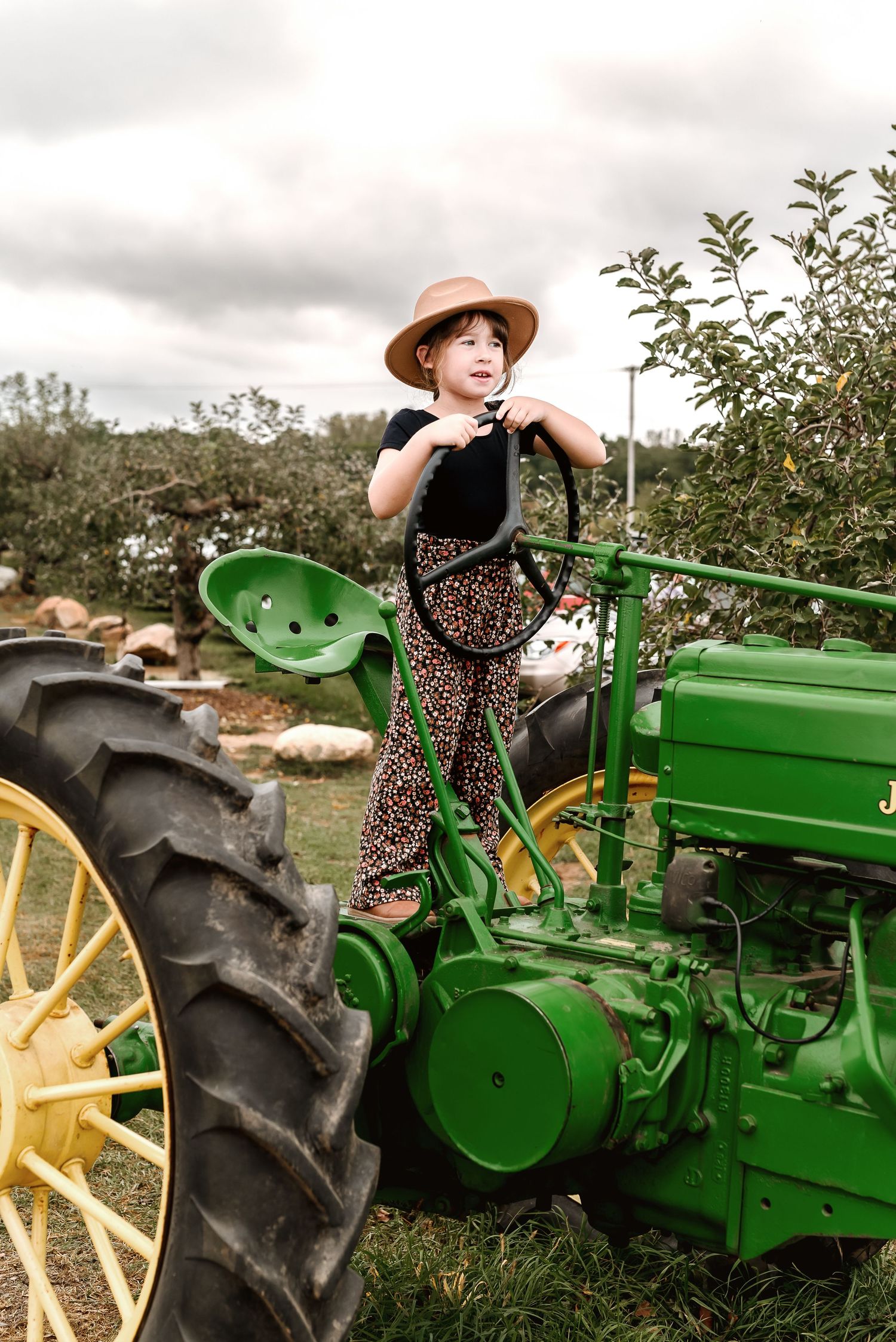 Someone in a brown hat sits atop a vintage green John Deere tractor with yellow wheels in an orchard setting.
