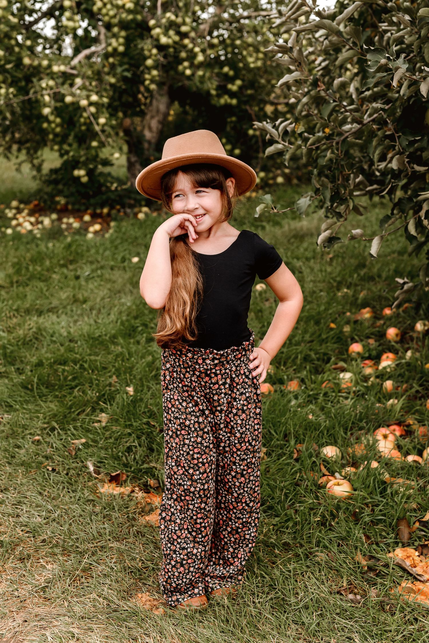 A person in a black shirt and leopard print skirt stands in a grassy orchard with scattered pumpkins on the ground.