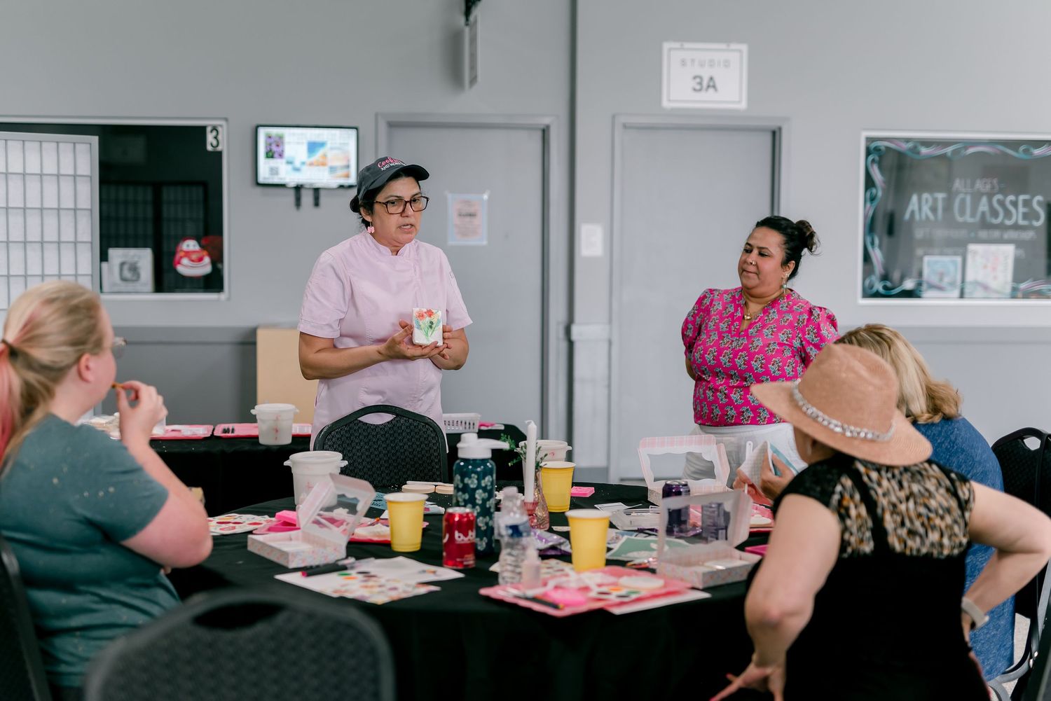 Women gathered around a table during an art class, with an instructor presenting in a bright community studio.