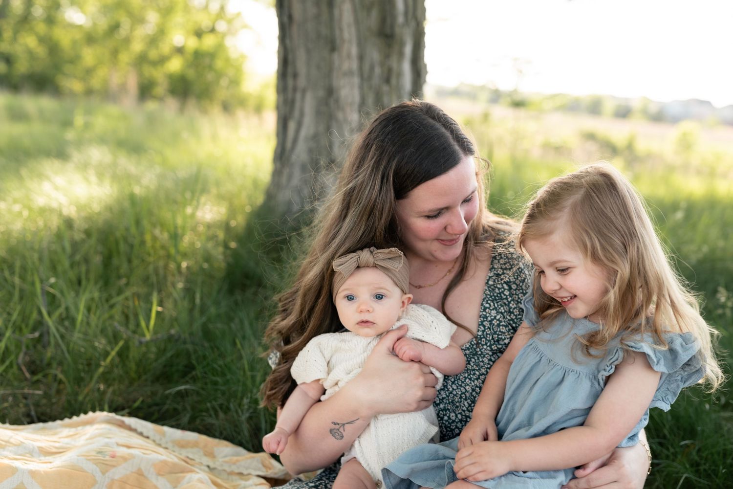 A mother sits under a tree in a green field, lovingly holding her baby and young daughter in a warm outdoor portrait.