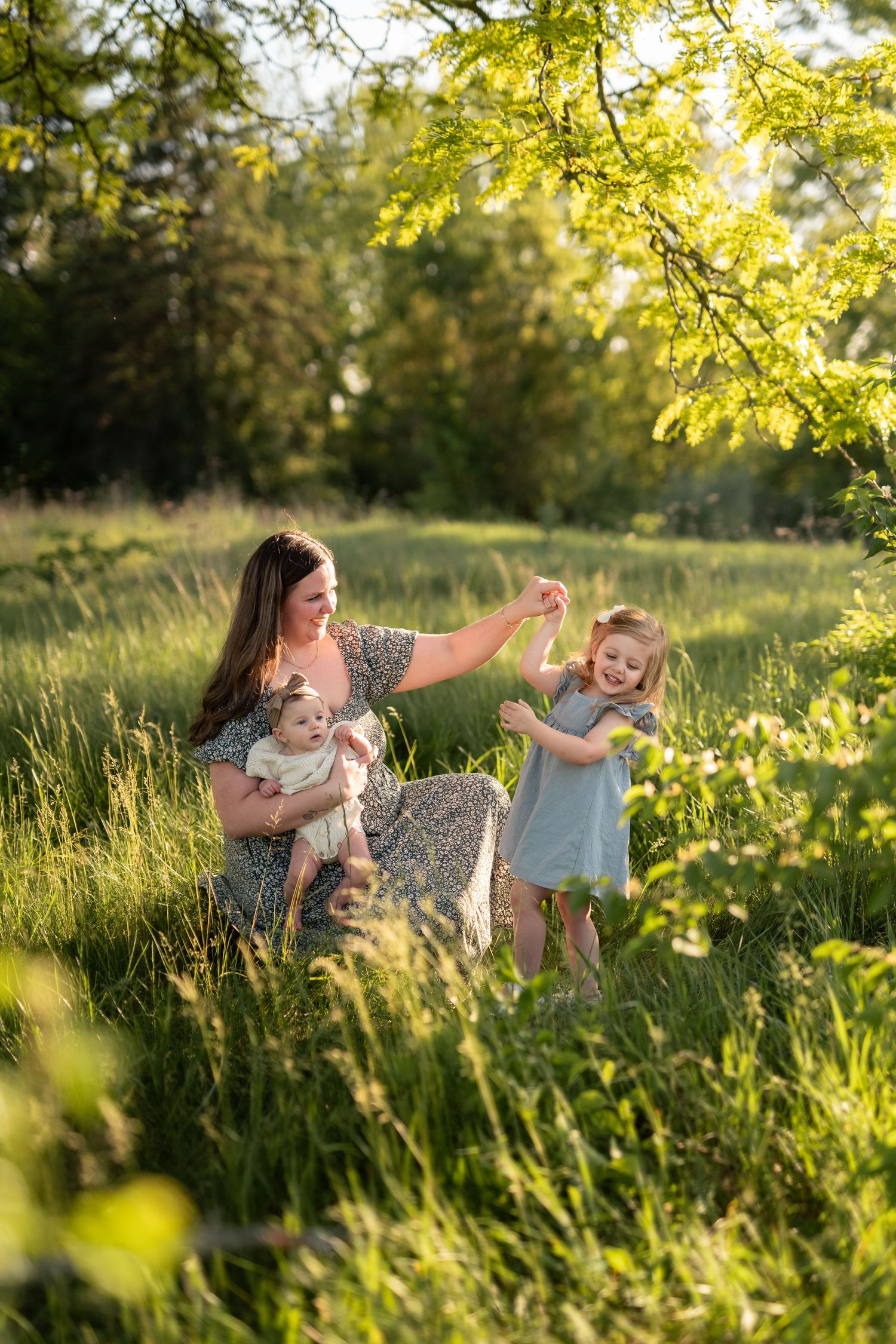 Mother holding baby while playing with toddler daughter in sunlit green meadow during golden hour.