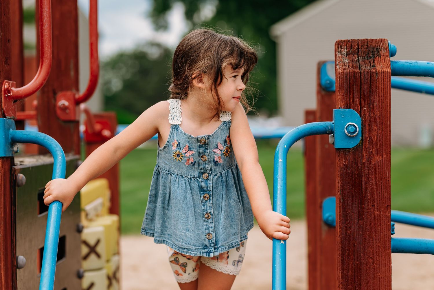 A child plays on a colorful playground structure with red and blue metal components on a summer day.