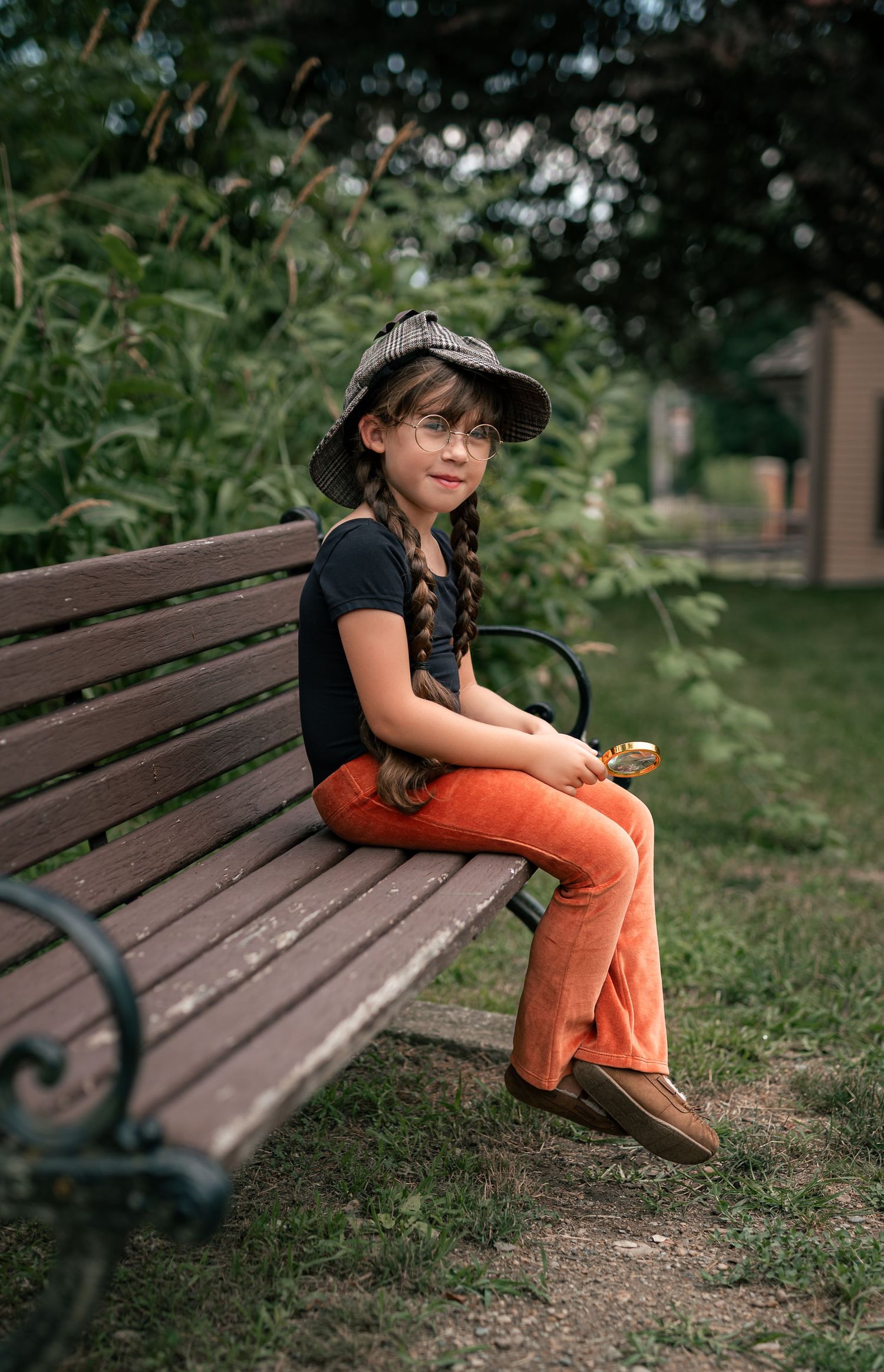 A young girl in orange pants, black tee, plaid cap, and glasses sits on a park bench holding a magnifying glass.