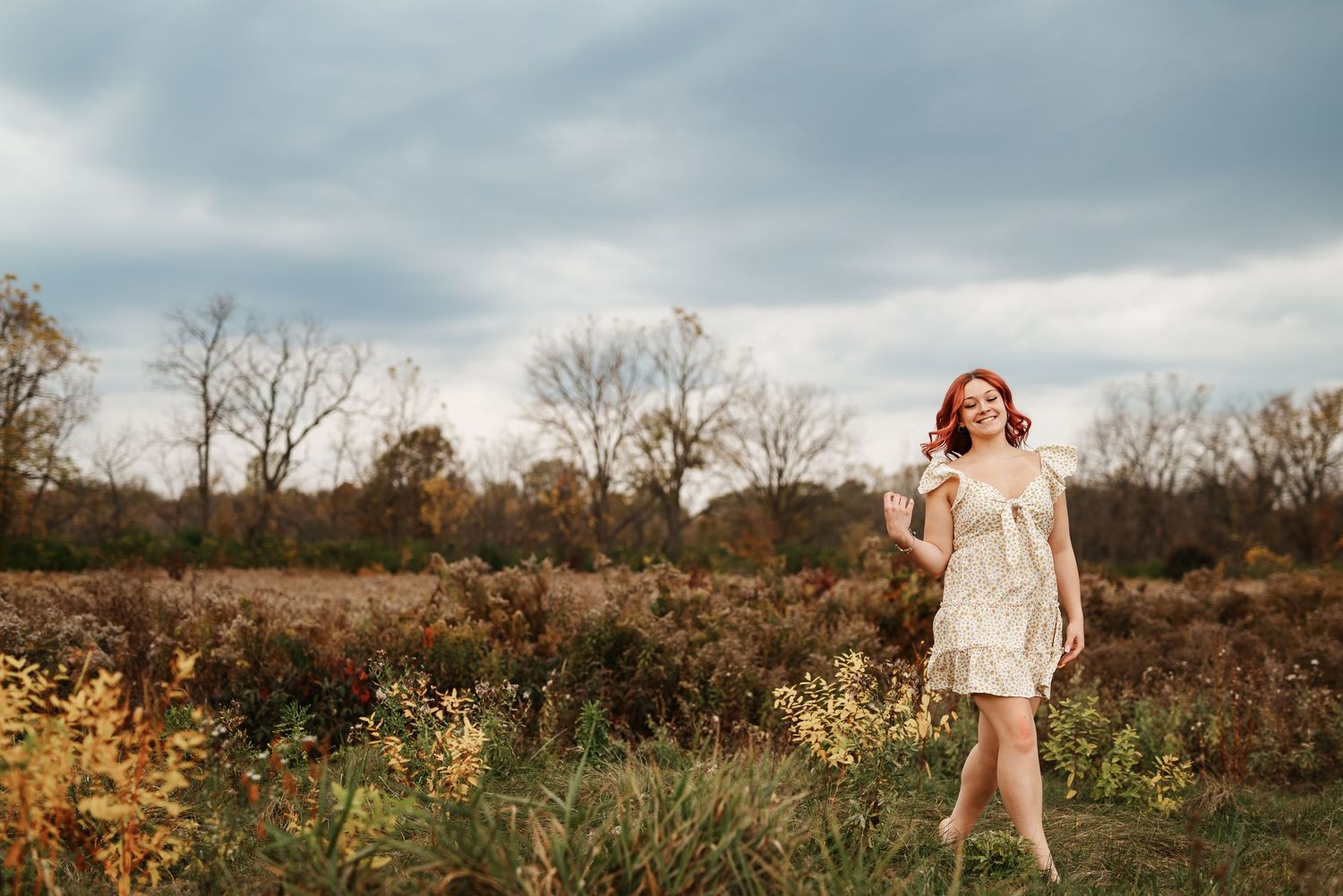 Red-haired woman in cream mini dress posing in golden autumn meadow under cloudy sky.
