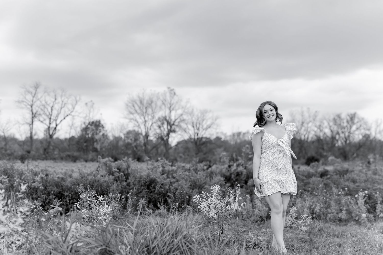 Black and white photo of a young woman in a light dress walking through a rural field on a cloudy day.