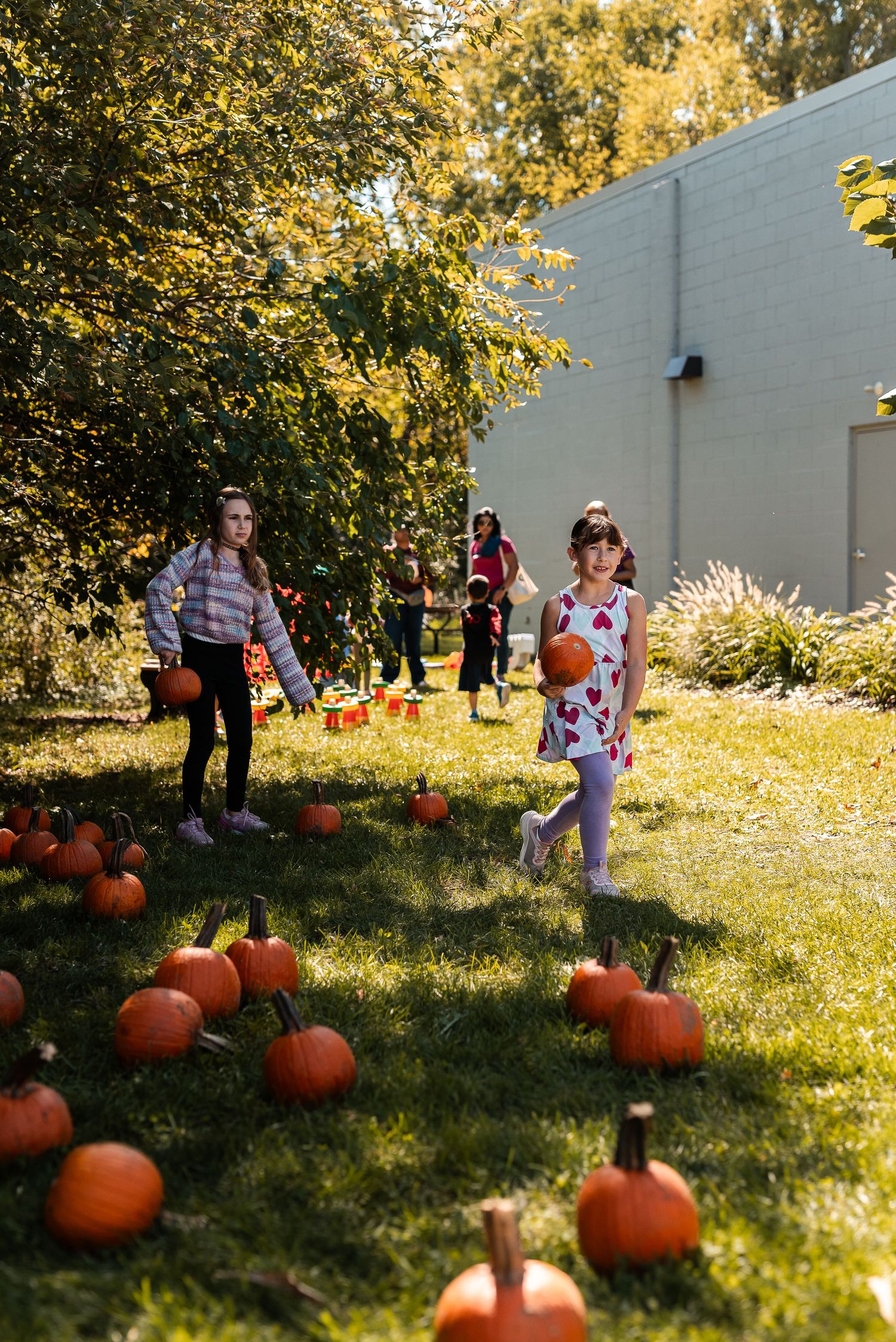 Two girls picking pumpkins from a sunny outdoor pumpkin patch at a fall festival event.