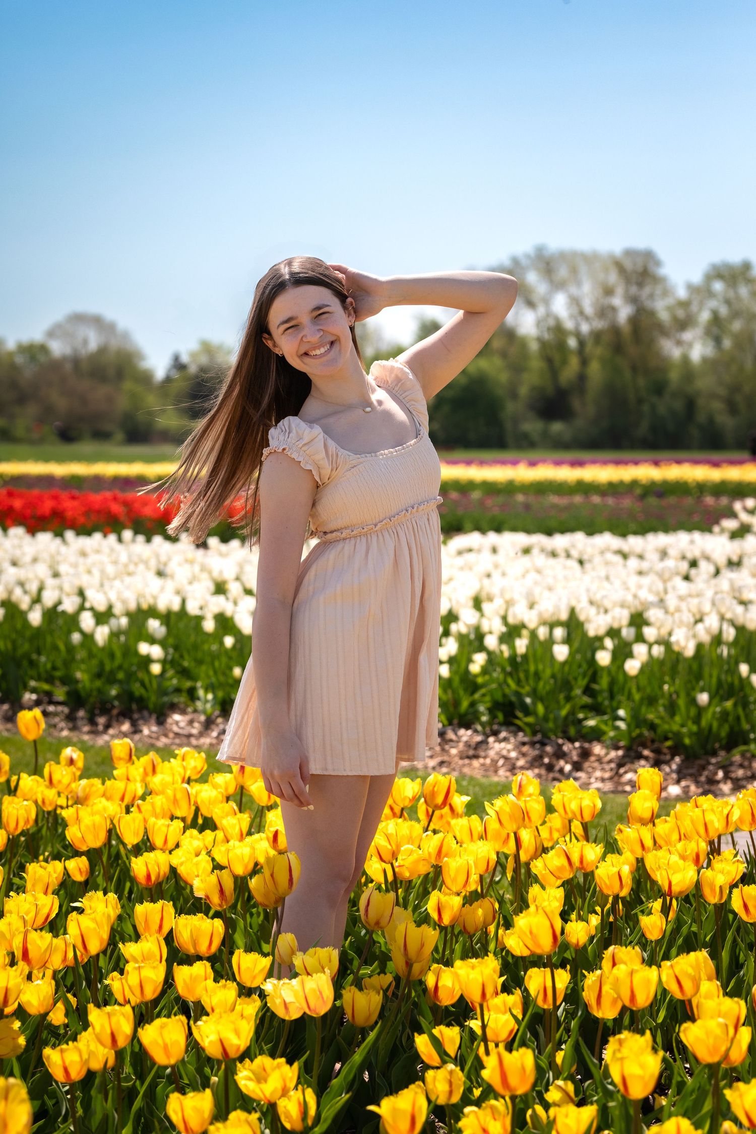 Smiling young woman in a beige dress standing among vibrant yellow tulips in a colorful flower field on a sunny day.