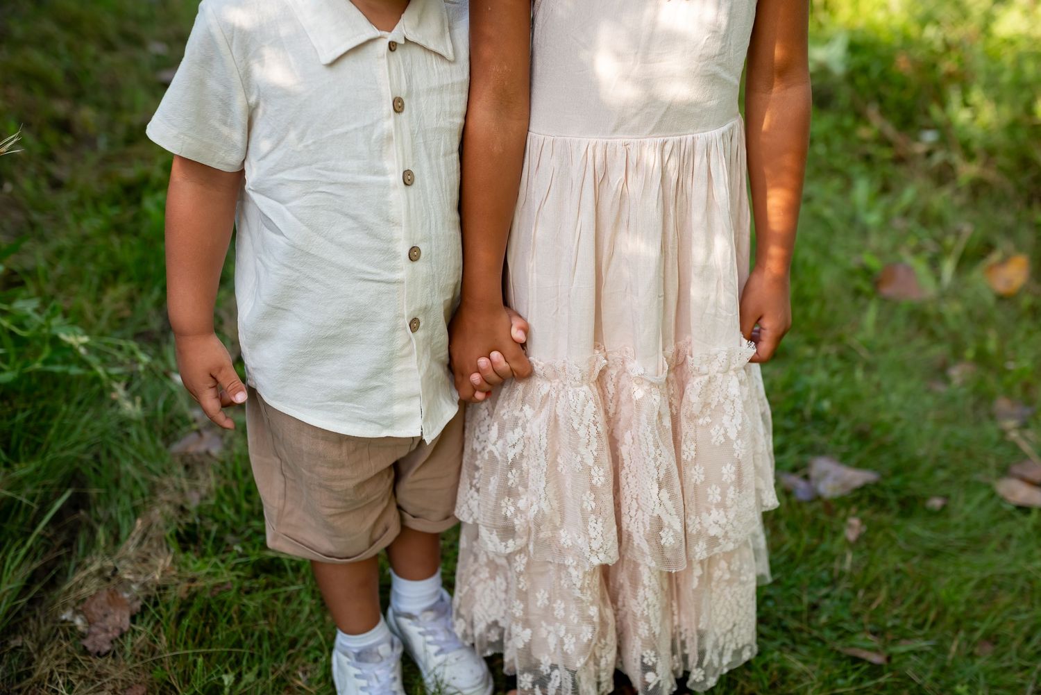 Two children holding hands outdoors, boy in white shirt and tan shorts, girl in white lace dress, standing on green grass.
