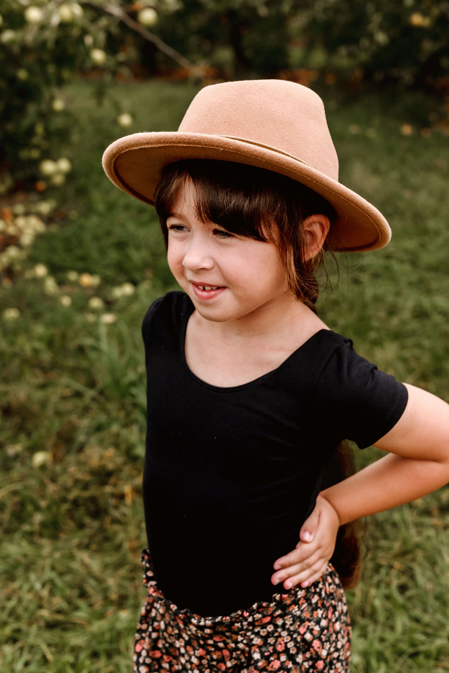 A child wearing a tan fedora hat, black shirt and floral skirt poses playfully outdoors against a green background.