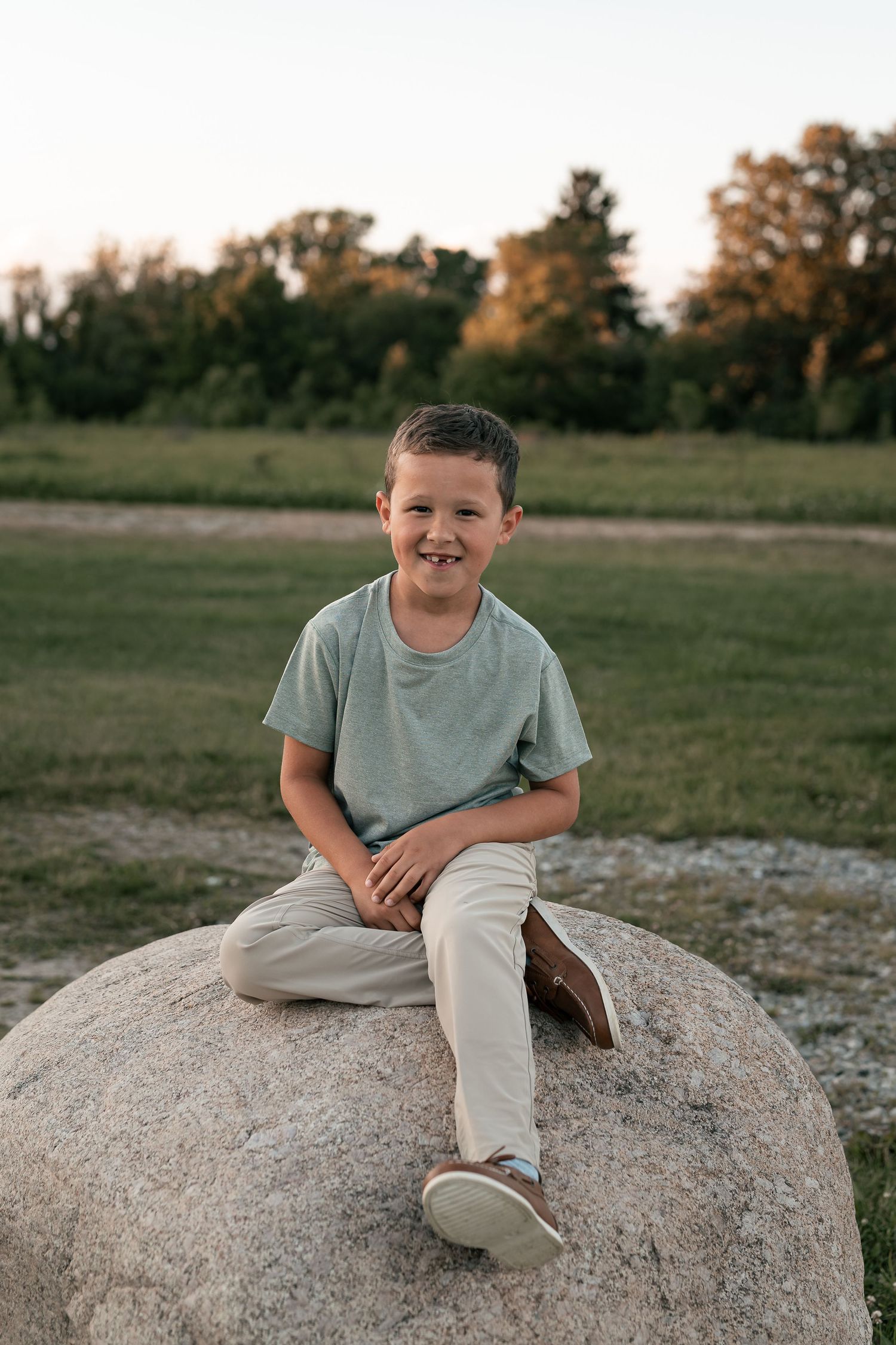 A smiling boy sits on a large rock in a green field at sunset, wearing a light blue t-shirt and beige pants.