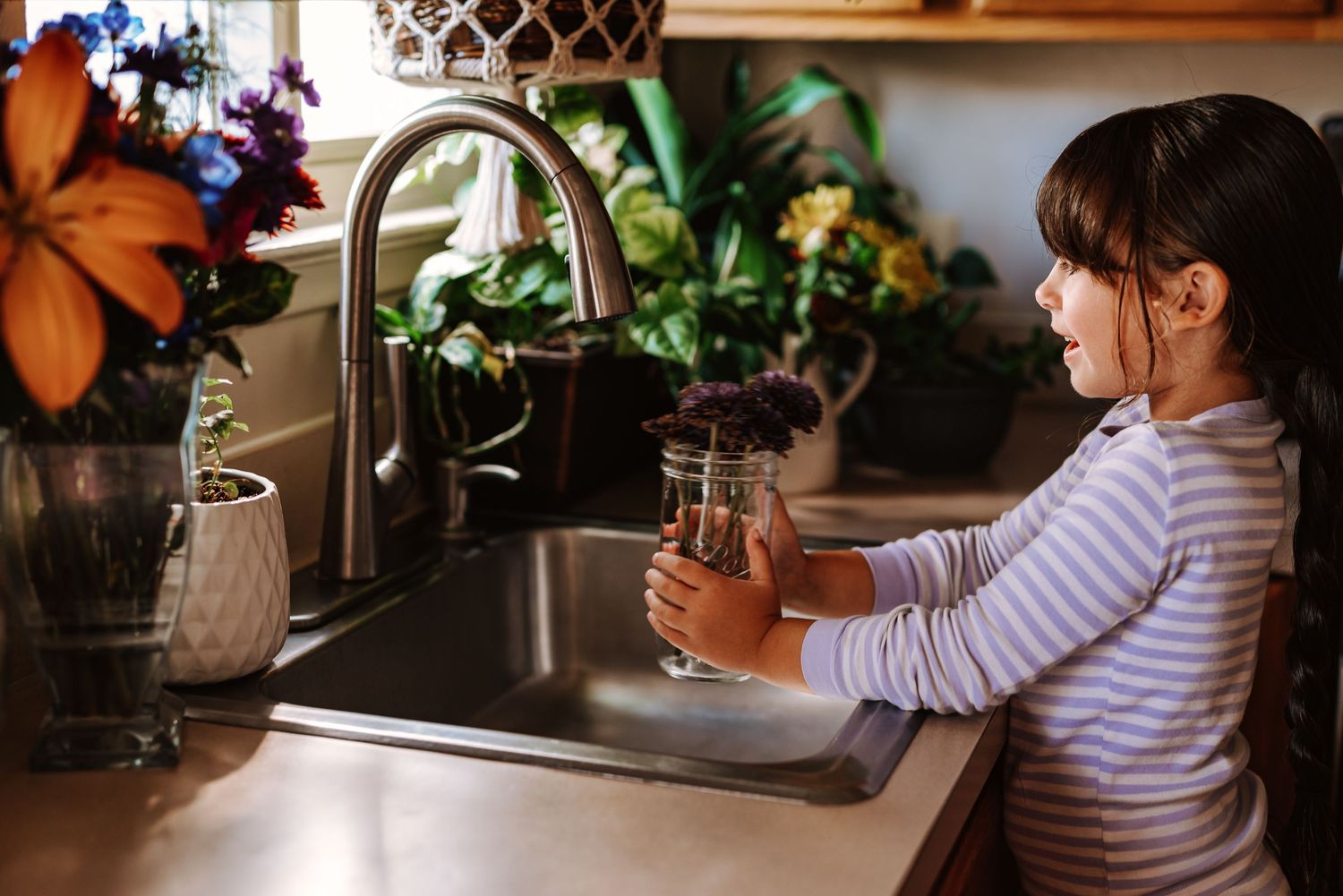 Small child in striped shirt washing dishes at kitchen sink with plants and flowers on counter.