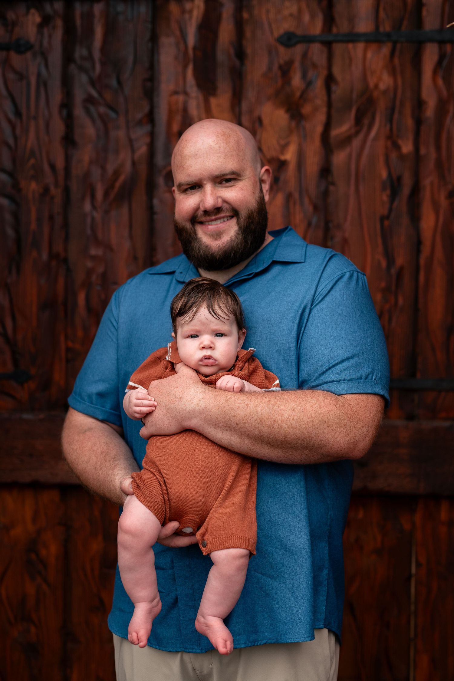 Smiling bald bearded man in blue shirt holding a newborn baby in a rust orange outfit before a wooden backdrop.