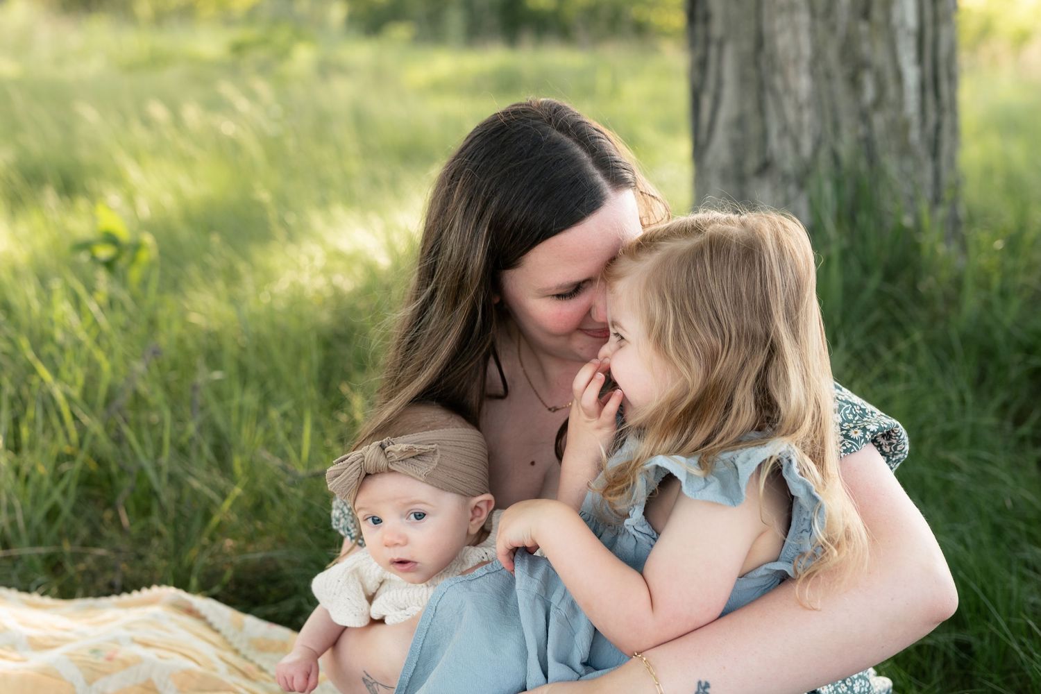 A mother tenderly kisses her toddler daughter while holding her infant baby outdoors in a lush green field.