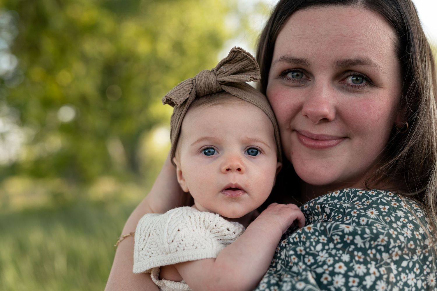 A young mother with brown hair holds her blue-eyed baby girl wearing a bow headband outdoors.