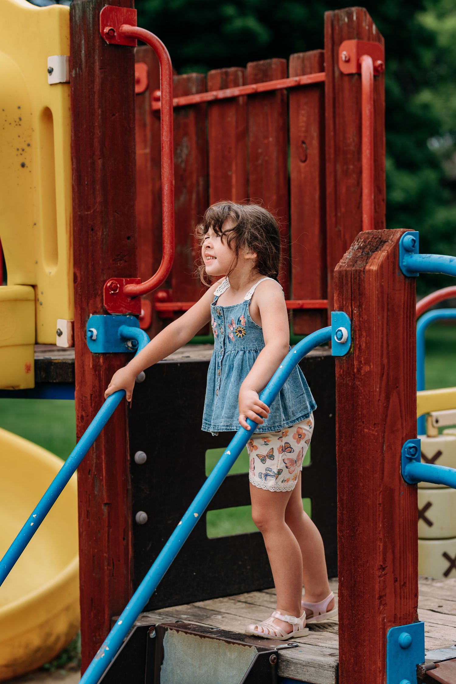 Child in denim dress plays on colorful playground equipment with red and blue climbing bars on a summer day.