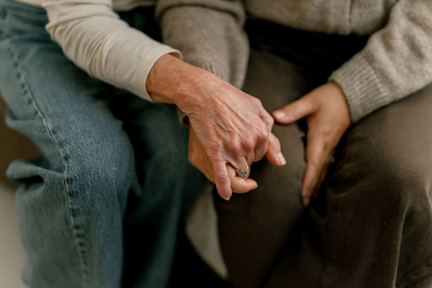 Two elderly people sitting side by side holding hands in a tender moment of comfort, love, and companionship.