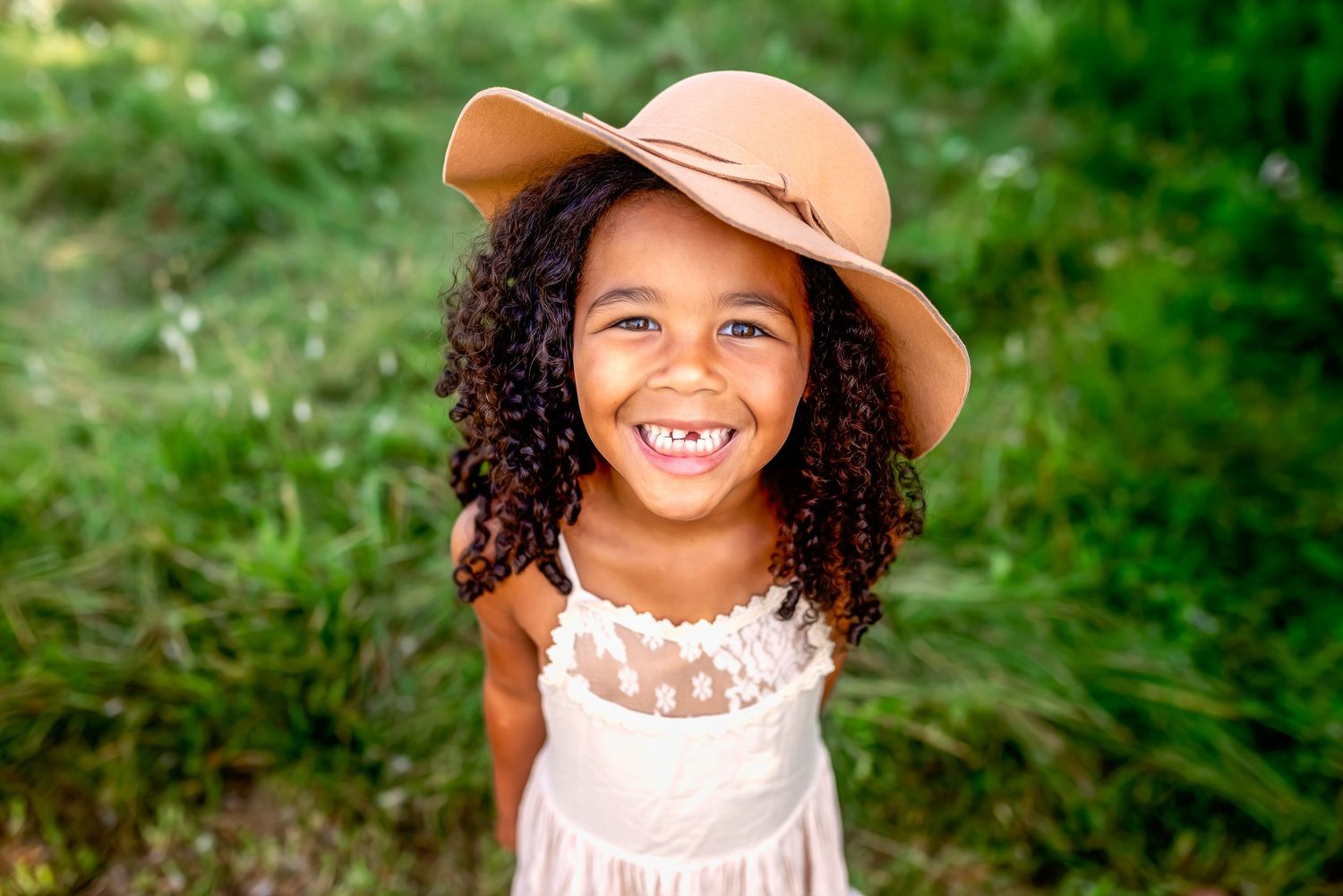 A young child with curly hair wearing a tan hat and white dress smiles brightly in a lush green outdoor setting.