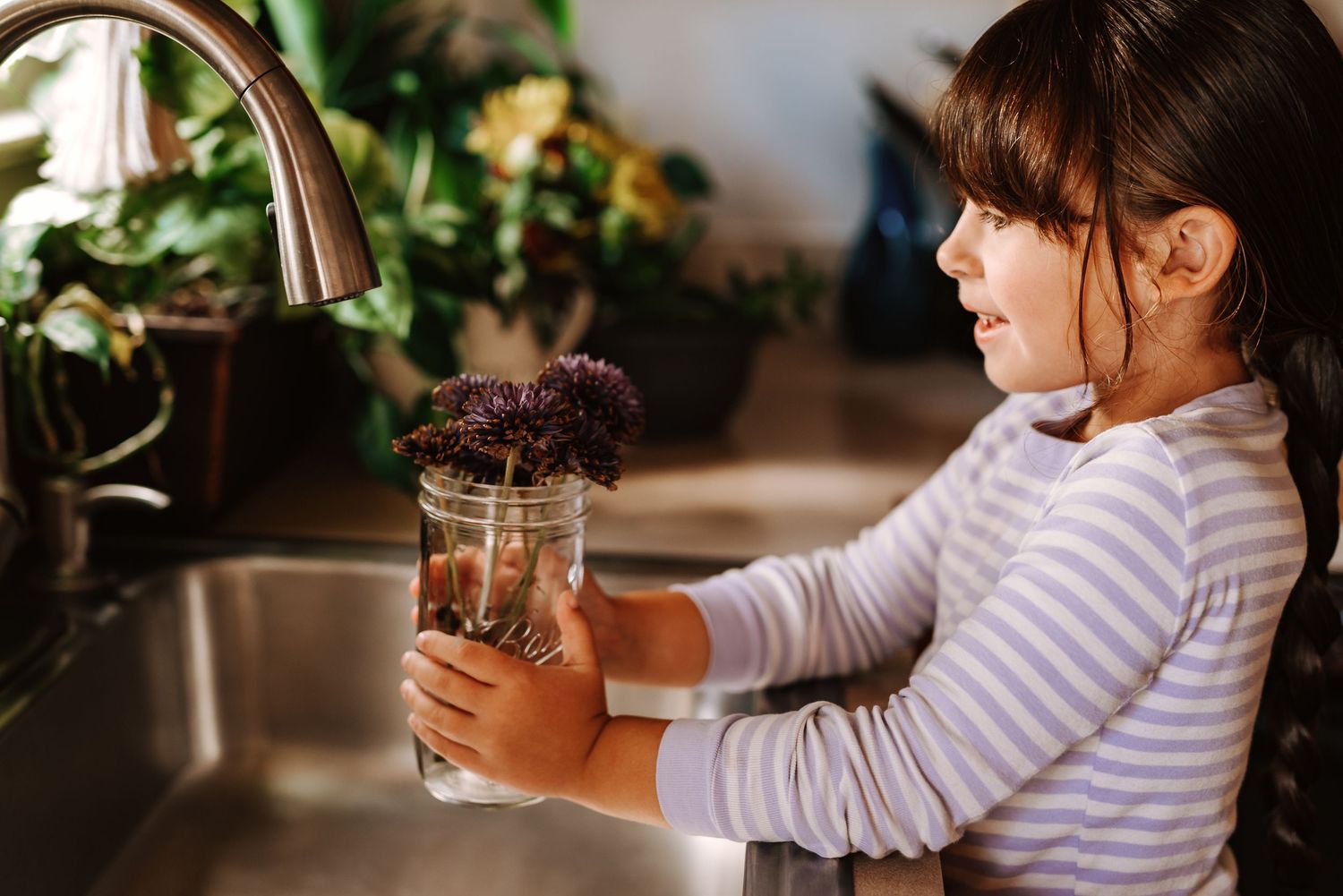 A person in a striped shirt washes a jar at a kitchen sink with plants in the background.