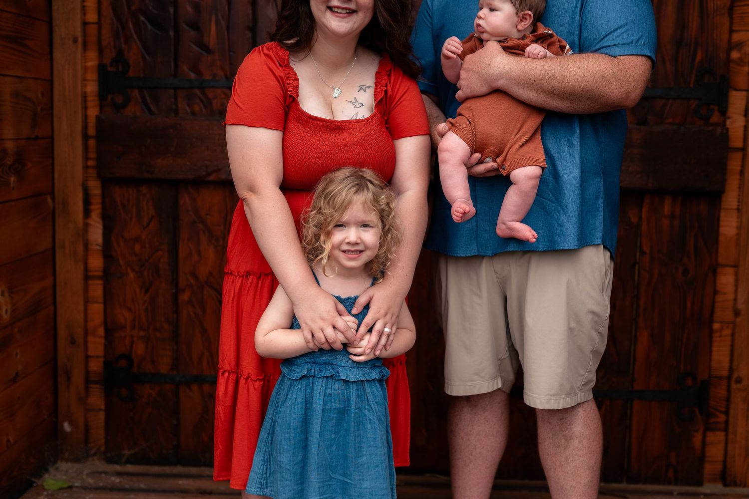 Family of four posing outdoors, mother in red dress, father in blue shirt holding newborn, young girl in blue dress smiling.