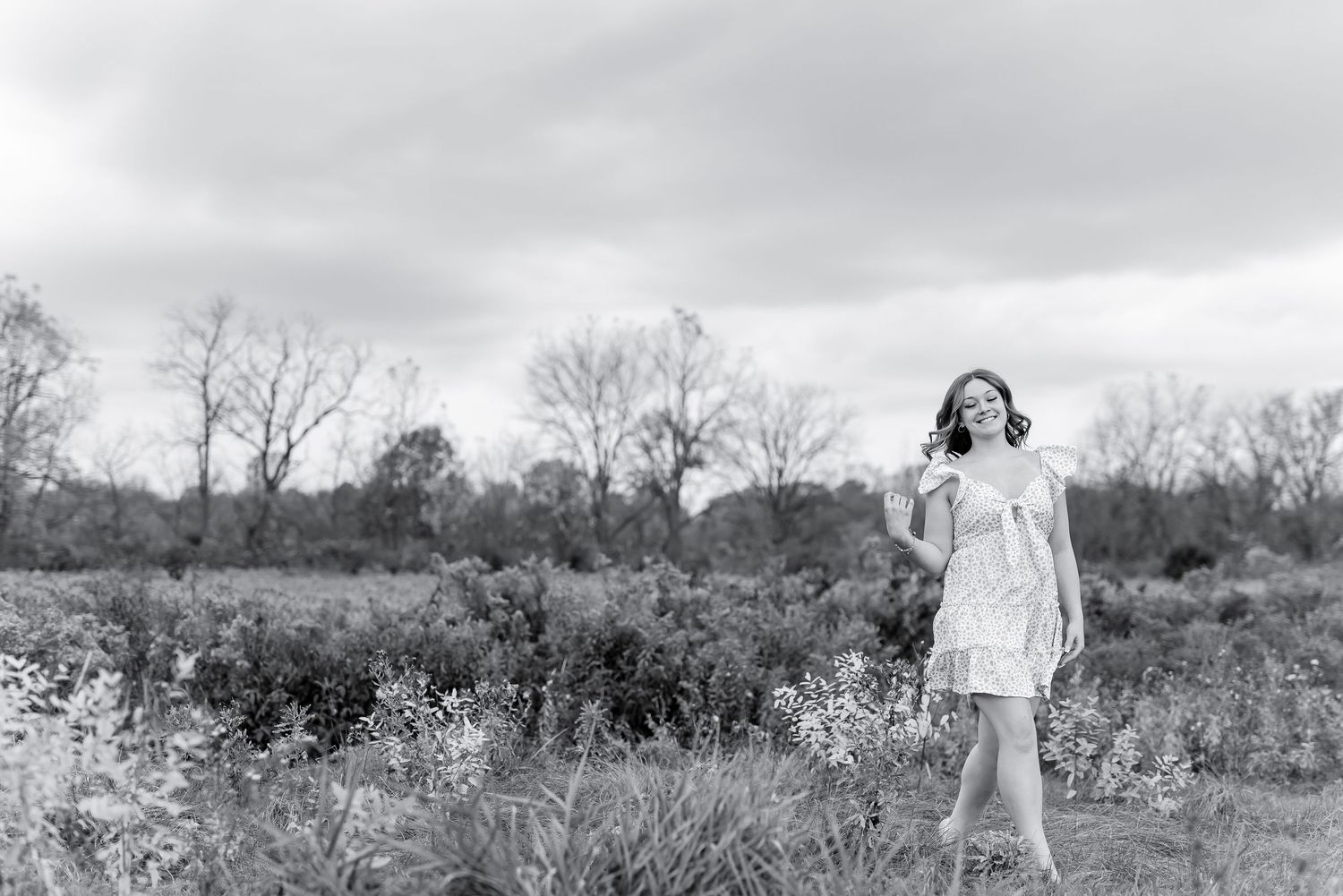 Black and white photo of young woman in flowy dress standing in open field on cloudy day.