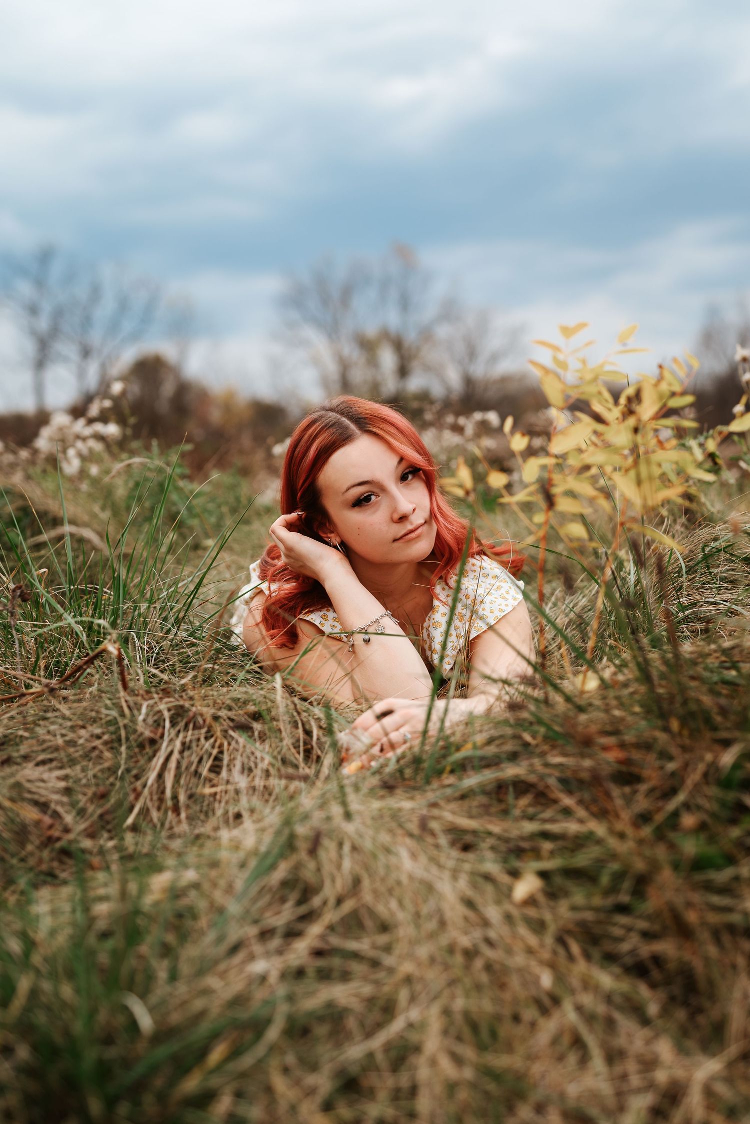 Red-haired young woman lying in a field of tall grass under a cloudy sky.