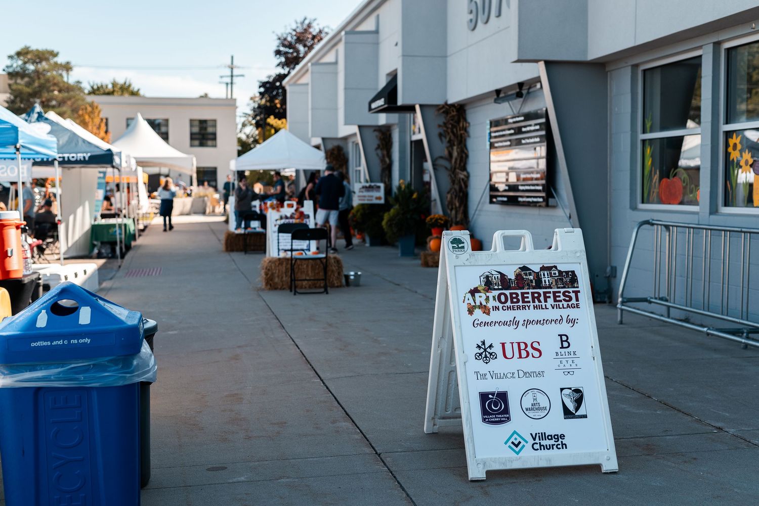 Oktoberfest community event with vendor booths and signage along a sunny outdoor shopping plaza walkway.
