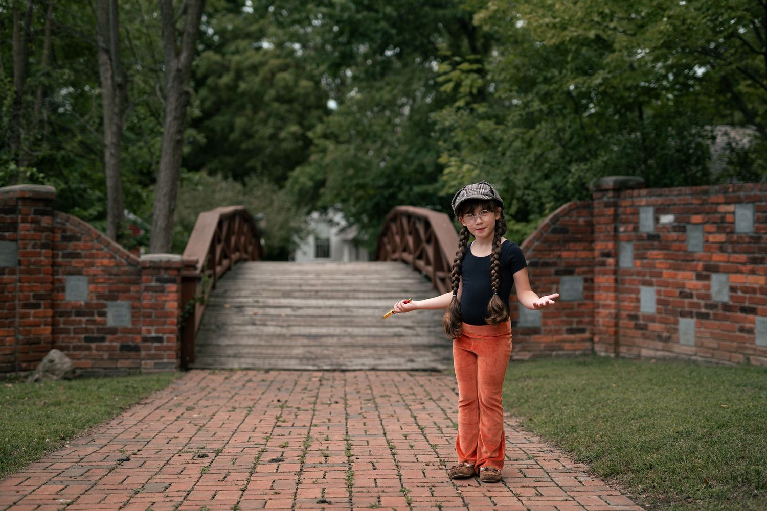 A young girl in orange pants and a black hat poses happily on a brick path near a stone bridge in a park.