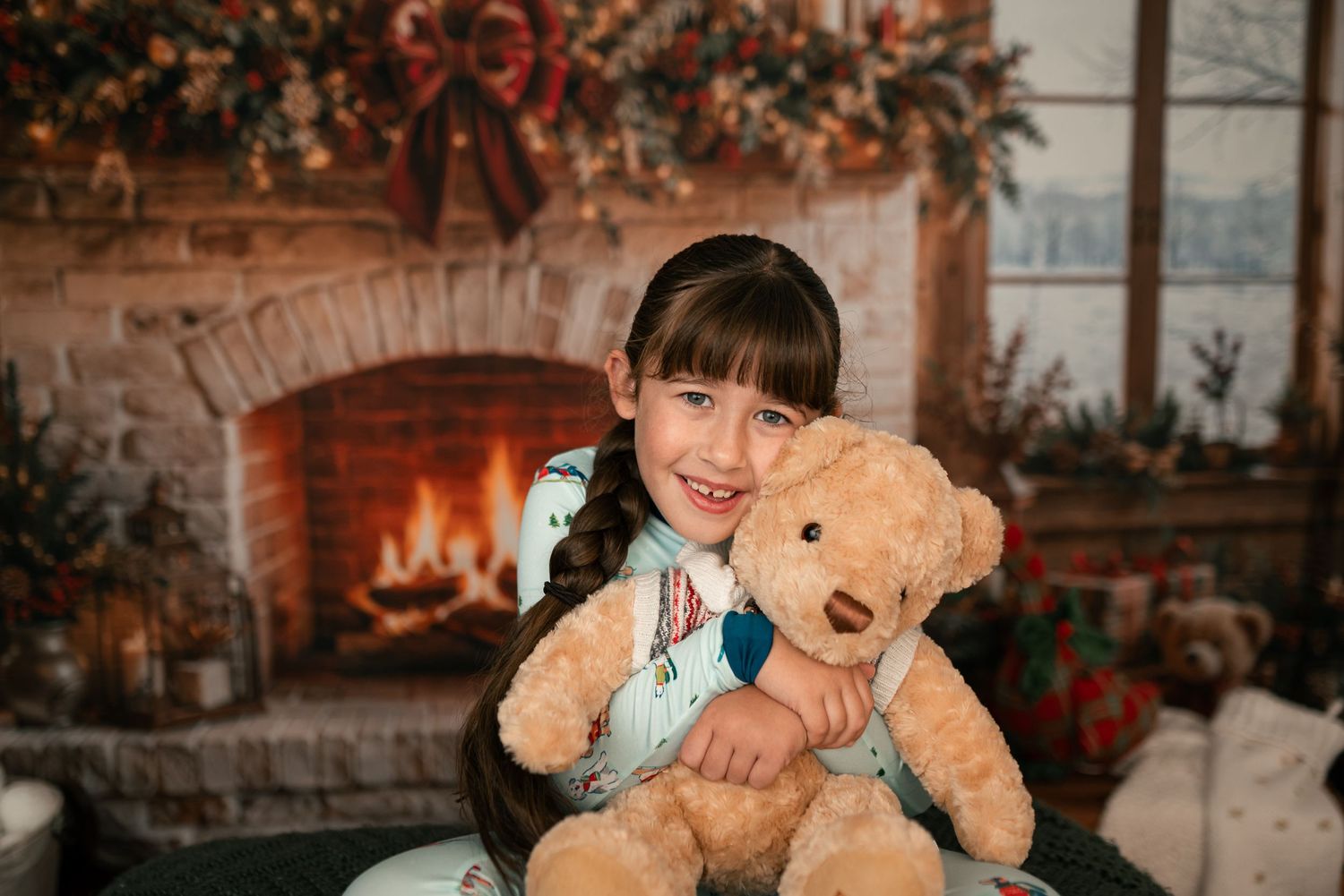 Smiling girl with long braided hair hugging a large teddy bear in front of a cozy brick fireplace decorated with a festive holiday garland.